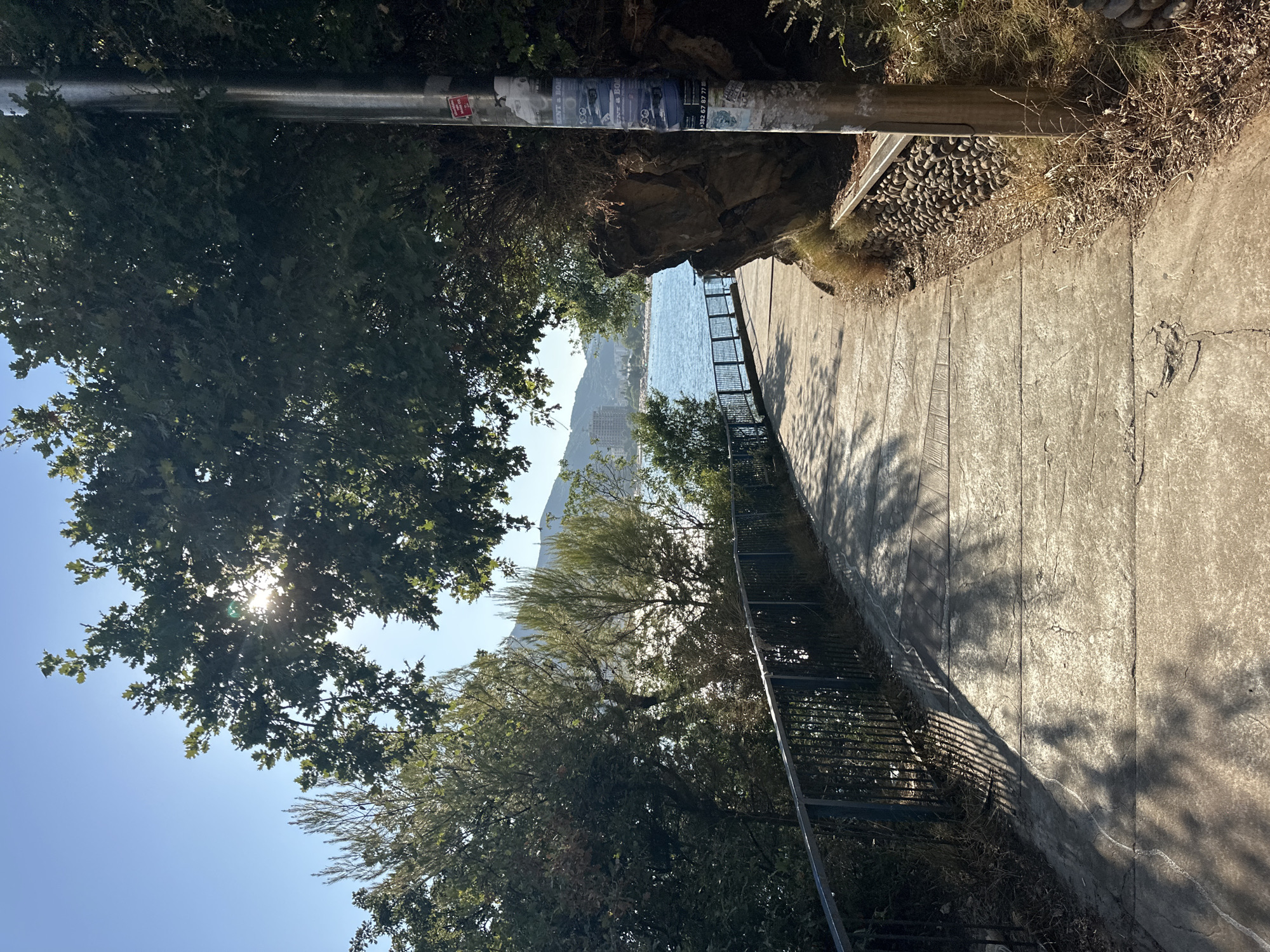 A paved pathway curves to the right, bordered by a black metal railing on the left and a rocky embankment on the right. The pathway is illuminated by sunlight filtering through trees, casting dappled shadows. To the left, beyond the railing, is a body of water with a town or city in the distance. A grey metal pole with several posters affixed to it stands on the right side of the path. The sky is clear and blue. This scene is likely along the promenade in Budva, Montenegro, a popular coastal town.