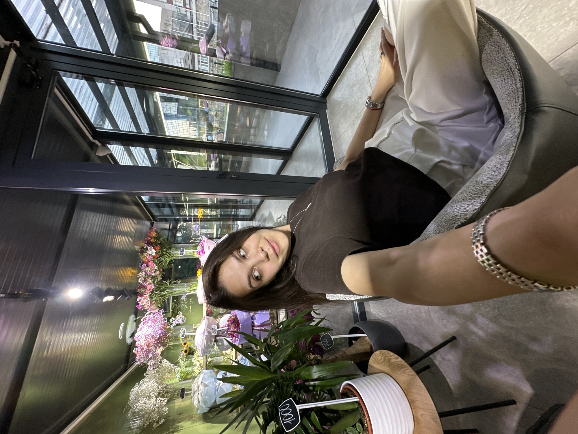 A young woman with long, dark hair is sitting in a chair in what appears to be a flower shop. She is wearing a dark brown t-shirt and light-colored pants. The flower shop has a modern design with large glass windows that allow natural light to enter. Various bouquets of flowers, in shades of pink, white, and purple, are displayed on shelves and in vases. A tall green plant is visible to the left of the woman, with price tags indicating "1000" and "700". The floor is tiled. Outside the window, buildings and a street scene are visible, suggesting an urban environment. The overall impression is that of a contemporary retail space. The location is identified as Chișinău, Moldova.