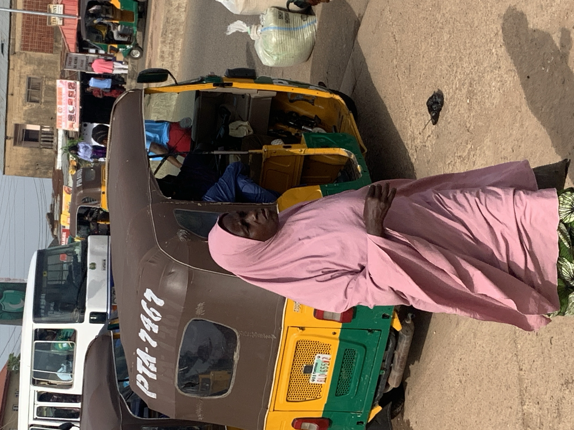 In the city of Gura Top, Nigeria, a woman wearing a light pink hijab and a patterned skirt stands in the foreground, partially obscuring a yellow and green auto-rickshaw. The auto-rickshaw has the license plate "BLO-655V2" and the marking "PTA-7467" on its brown roof. To the left of the auto-rickshaw, the front of a white bus is visible. In the background, buildings line the street, and other auto-rickshaws and people are present, suggesting a busy marketplace or transportation hub. A large white sack is visible to the right of the auto-rickshaw, and a dark object lies on the ground in the lower right corner. The scene appears to be outdoors on a paved surface.