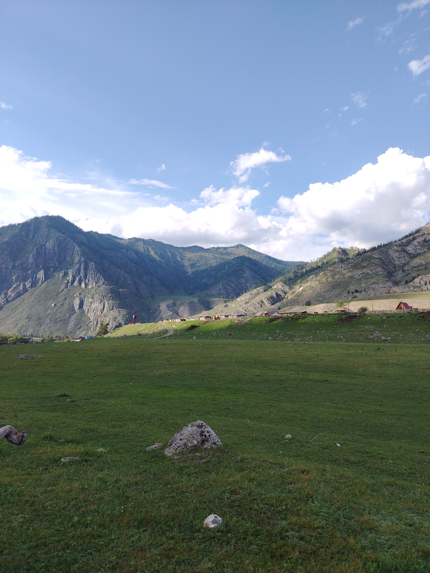 The image depicts a rural landscape in Iodro, Russia, under a partly cloudy sky. In the foreground, a vast expanse of green grass is dotted with several rocks, one notably larger than the others. The middle ground features a gentle slope leading up to a settlement of small houses with red roofs, arranged along a curve. A dirt road winds through the settlement. Behind the houses, a darker, steeper mountain range rises, its slopes partially covered in vegetation and marked by rocky outcrops. The sky above is a pale blue, with scattered white clouds. The overall impression is of a serene, natural environment with a small human settlement integrated into the landscape.
