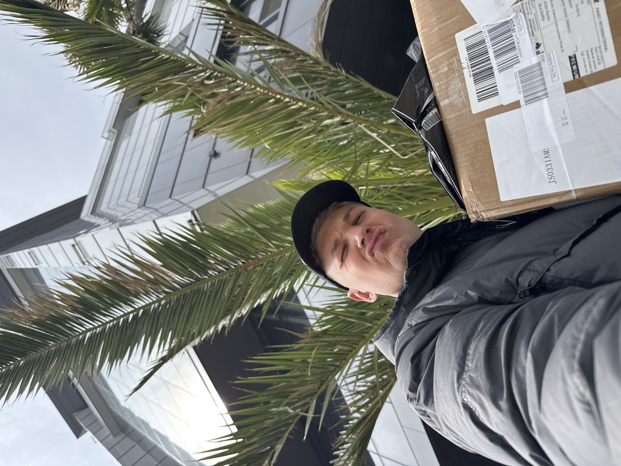 A man in a black baseball cap and gray puffy jacket is holding a cardboard package against his body, with the labels facing the camera. The labels indicate the package is international and has a return address of 4004 Plovdiv, Bulgaria. He is standing outside, as evidenced by the palm tree fronds in the background, and a modern building with a glass facade is visible in the upper left corner of the frame. The photo appears to be taken from a low angle, looking upwards.
