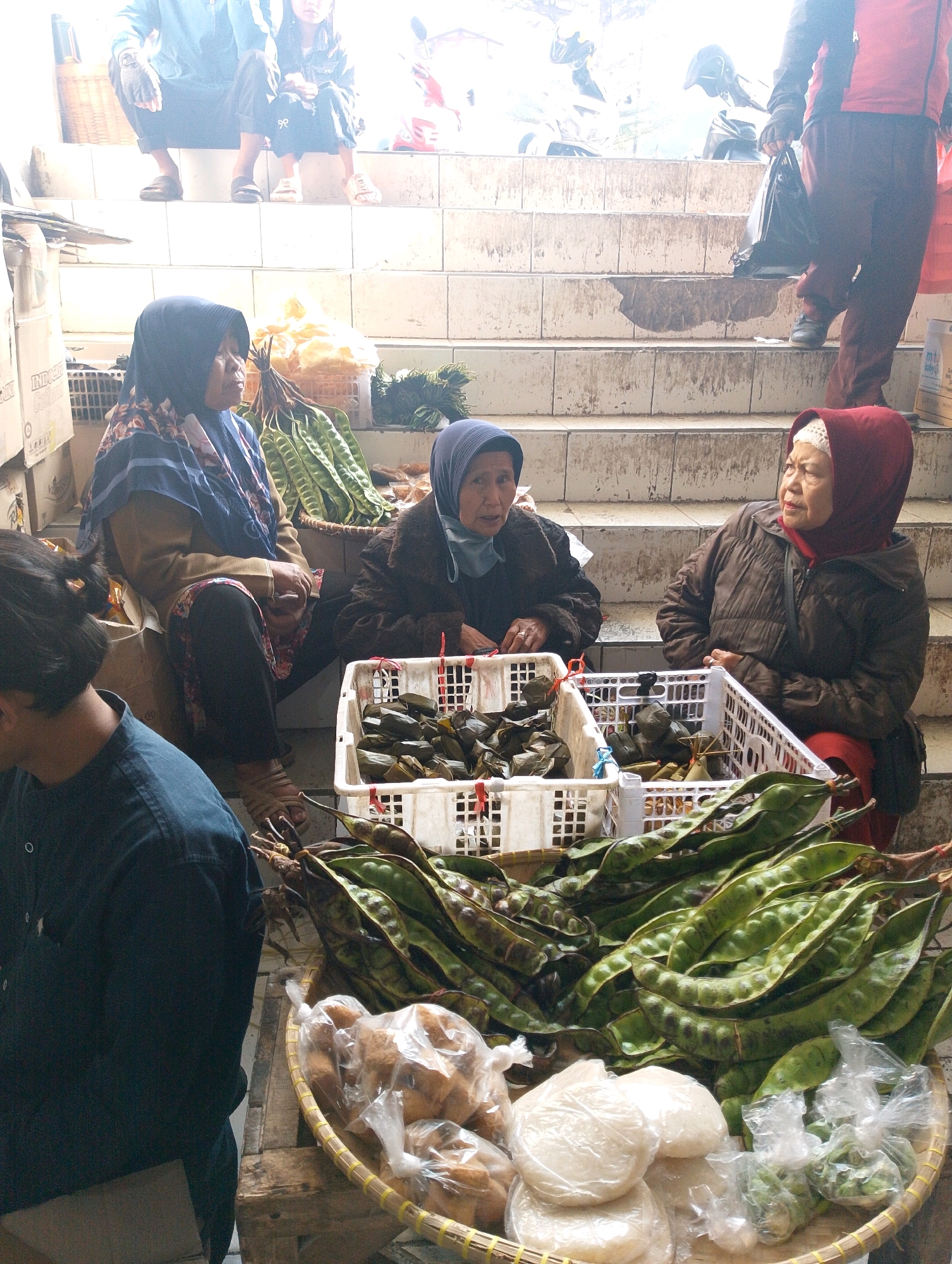 The image depicts a street market scene in Lembang, Indonesia. In the foreground, a person wearing a dark blue shirt is partially visible from the side. To their right, a woven basket overflows with long, green petai pods. Beside the basket are plastic-wrapped bundles of what appear to be food items, some round and white, others more amorphous.
Further back, three women are seated. The woman on the left wears a blue hijab and a patterned brown top. The woman in the center has a light blue hijab and is wearing a dark brown jacket. The woman on the right wears a red hijab and a similar brown jacket. In front of the seated women are white plastic crates containing more petai pods and wrapped food items.
Behind the seated individuals, white concrete stairs are visible, some of which are stained and appear damp. On the stairs, two people are partially visible; one wearing a red top and dark pants is walking away from the camera, holding a black plastic bag, and another person in a blue shirt is seated. In the background, the environment appears bright and outdoors, with hints of motorcycles and a red object that could be a bag or another vehicle. The overall impression is a bustling marketplace with vendors selling fresh produce and prepared food items.