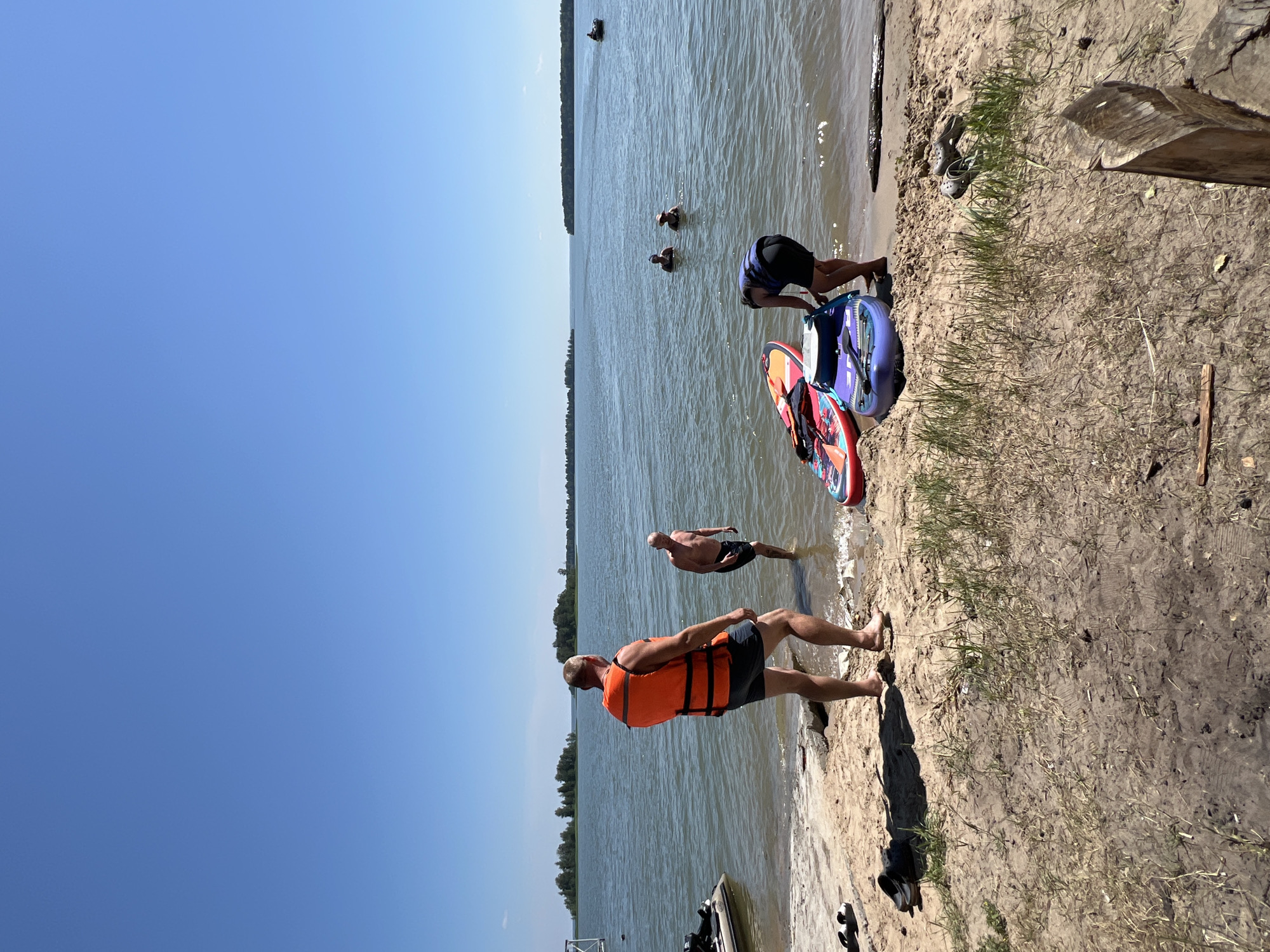 The image depicts a sunny day at a sandy beach on a body of water, likely a lake given the calm conditions and distant tree-lined shore. In the foreground, a man wearing an orange life vest and dark shorts stands facing away from the camera. To his right, another man, shirtless and wearing dark shorts, stands in the shallow water. Several paddleboards are present on the sand near the water's edge. Further into the water, two people are visible, and in the distance, a boat can be seen on the water. The environment is characterized by clear blue skies, calm water, and a sandy shoreline with sparse vegetation. The location is identified as the city of Borovoe, Russia.