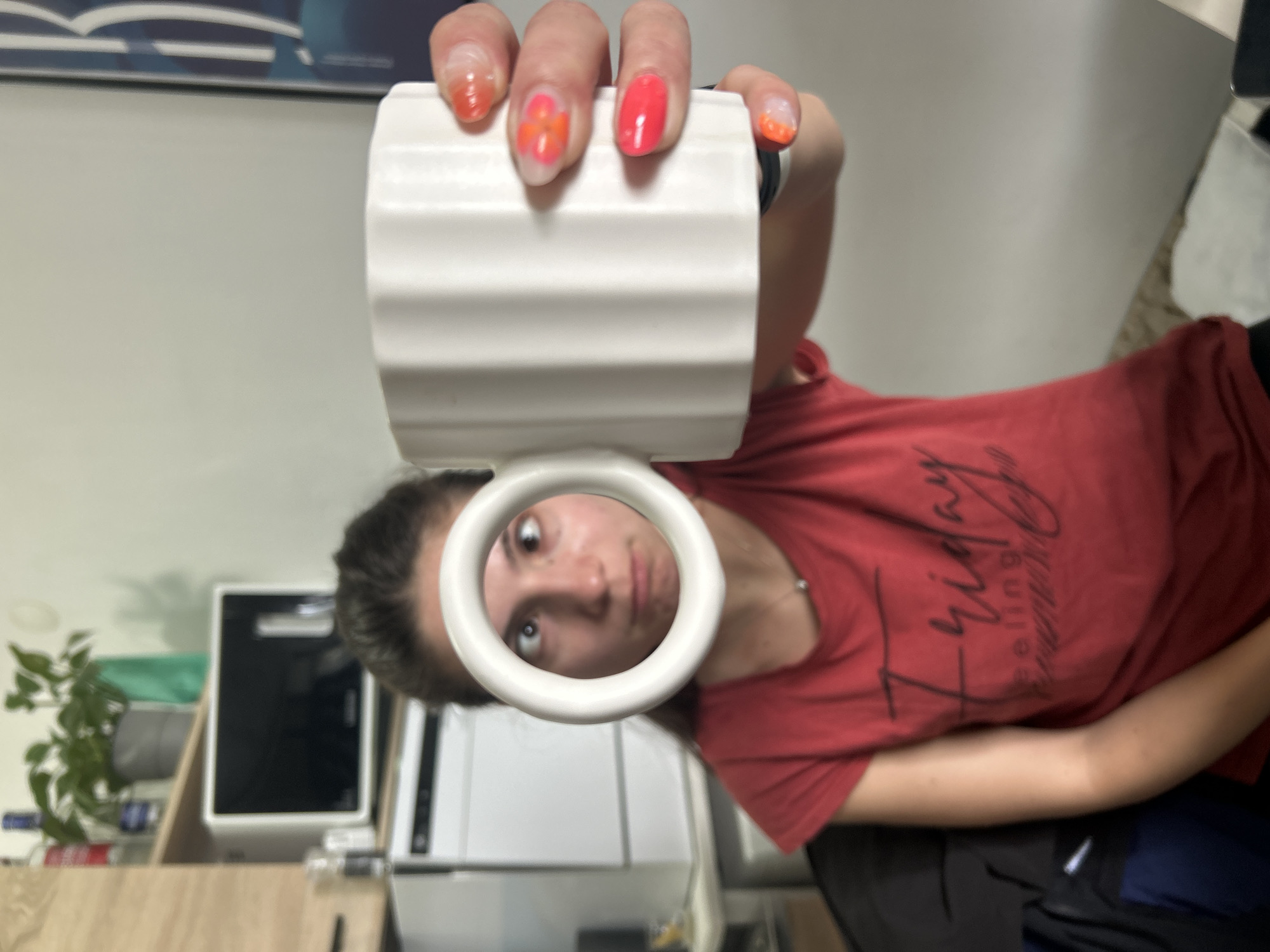 A woman is holding a white ceramic mug up to her face, peering through the handle. She is wearing a red t-shirt with black writing. Her fingernails are painted with various orange designs. Behind her is a desk with a black and white electronic device, and a small green plant. There is a white wall in the background. The image does not contain information about the location of the photo in the city of Sofia, Bulgaria.