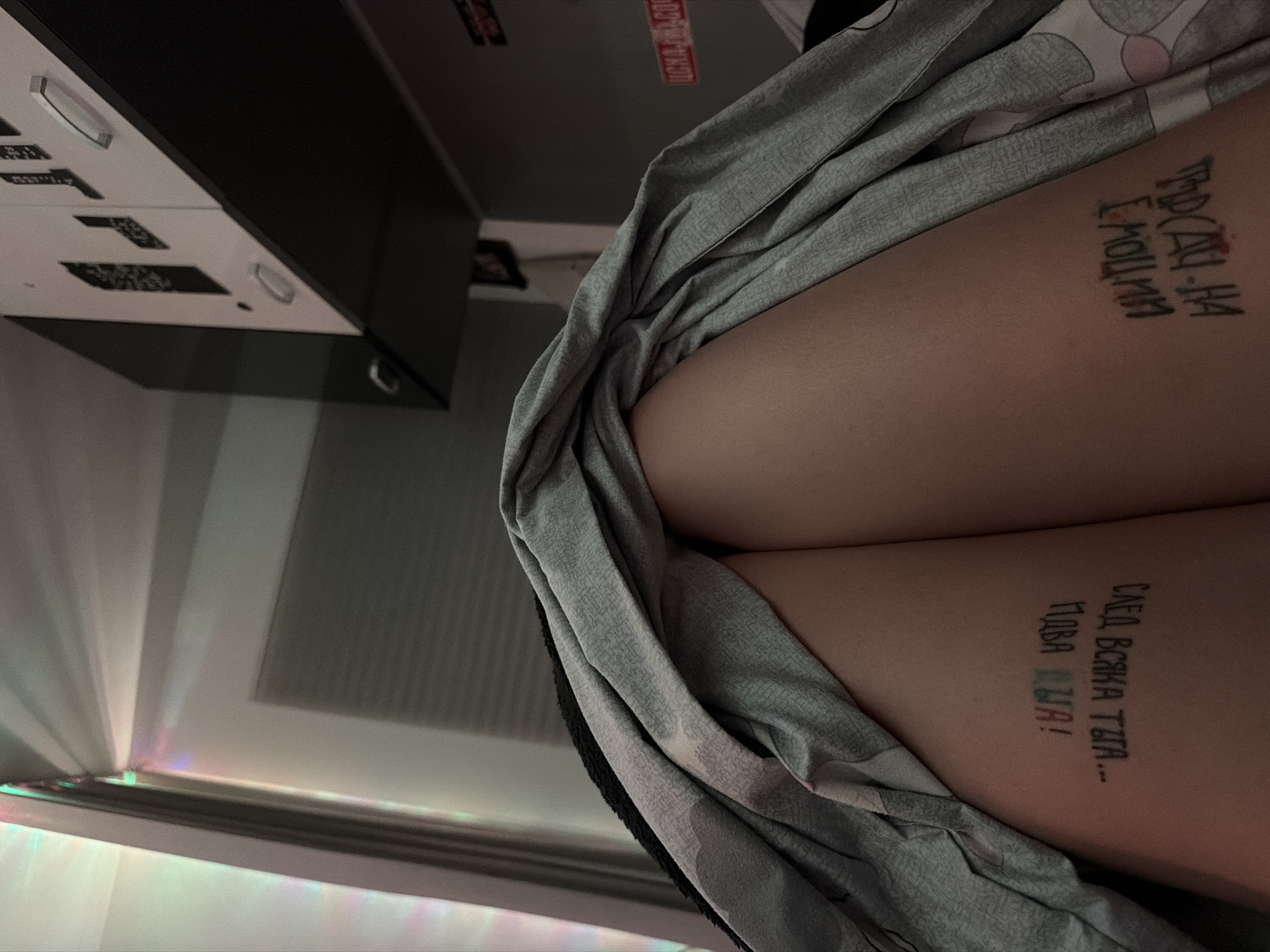 The image shows a person's thighs with Russian text tattoos, draped with a gray patterned fabric. In the background, there is a cabinet and a window with light shining through. The location of the image is unavailable.