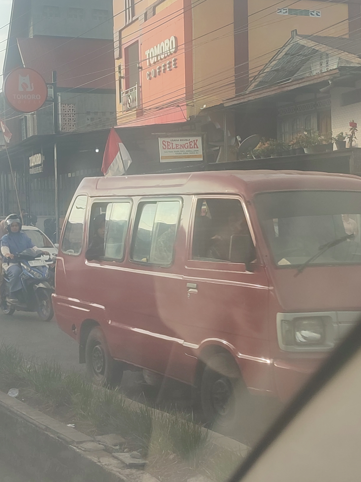 A red minivan is parked on a street in Adirejo, Indonesia. The minivan is facing the right side of the frame. The front of the minivan is visible. The minivan has two doors and a windshield. There are people sitting in the minivan. A woman on a scooter is riding next to the minivan. She is wearing a blue shirt and a helmet. There are buildings in the background. One building has a sign that says "TOMORO COFFEE". Another building has a sign that says "SELENGAK". There are also Indonesian flags visible. The street is paved and there are some plants on the side of the road. The sky is overcast.