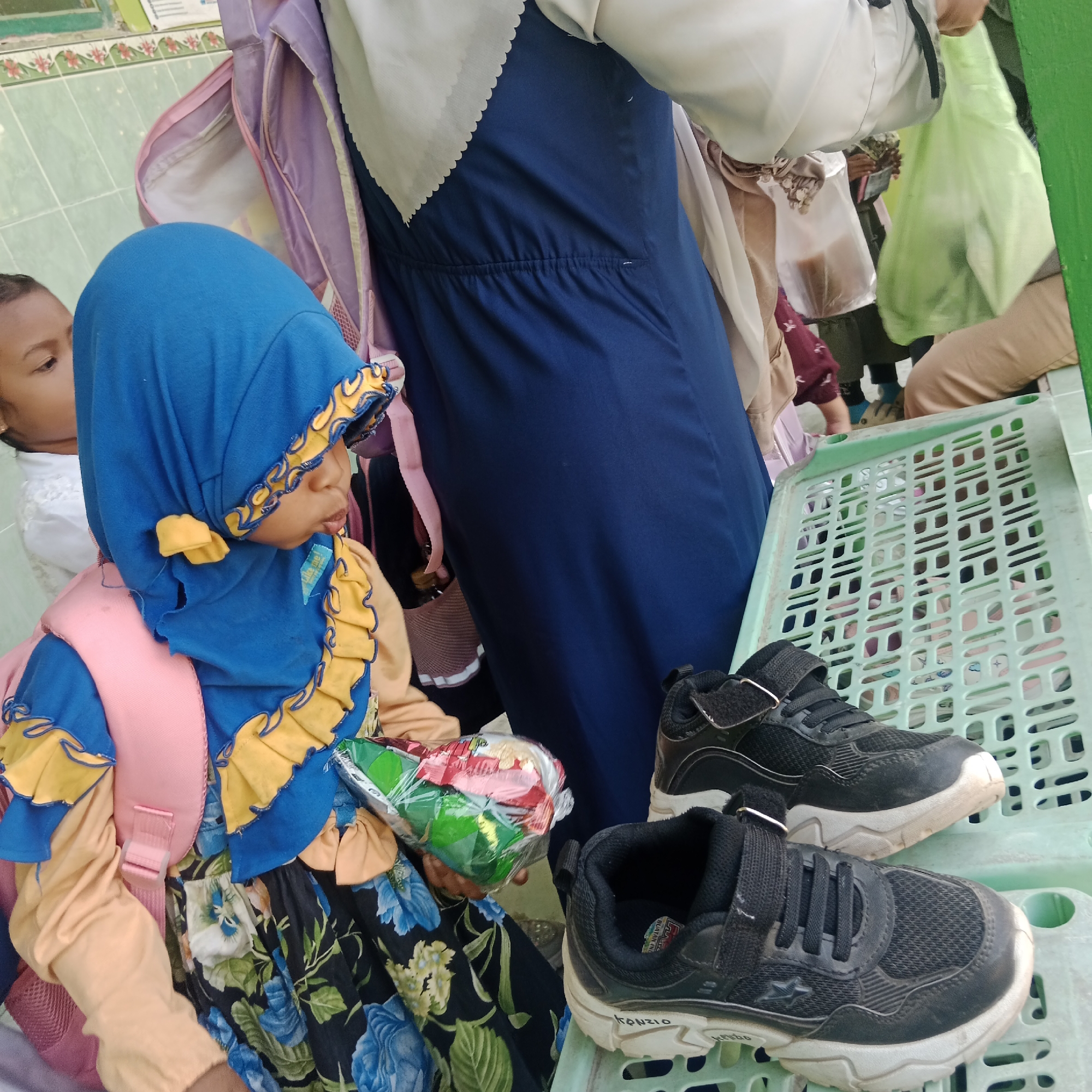 In Tegowanu Kulon, Indonesia, a young girl wearing a blue hijab with yellow trim and a floral-patterned dress is holding a packaged item. She has a pink backpack on. To her right, two pairs of black sneakers with white soles are placed on a light green plastic basket. In the background, several people are visible, some carrying bags. The environment appears to be outdoors, with a tiled wall in the background.