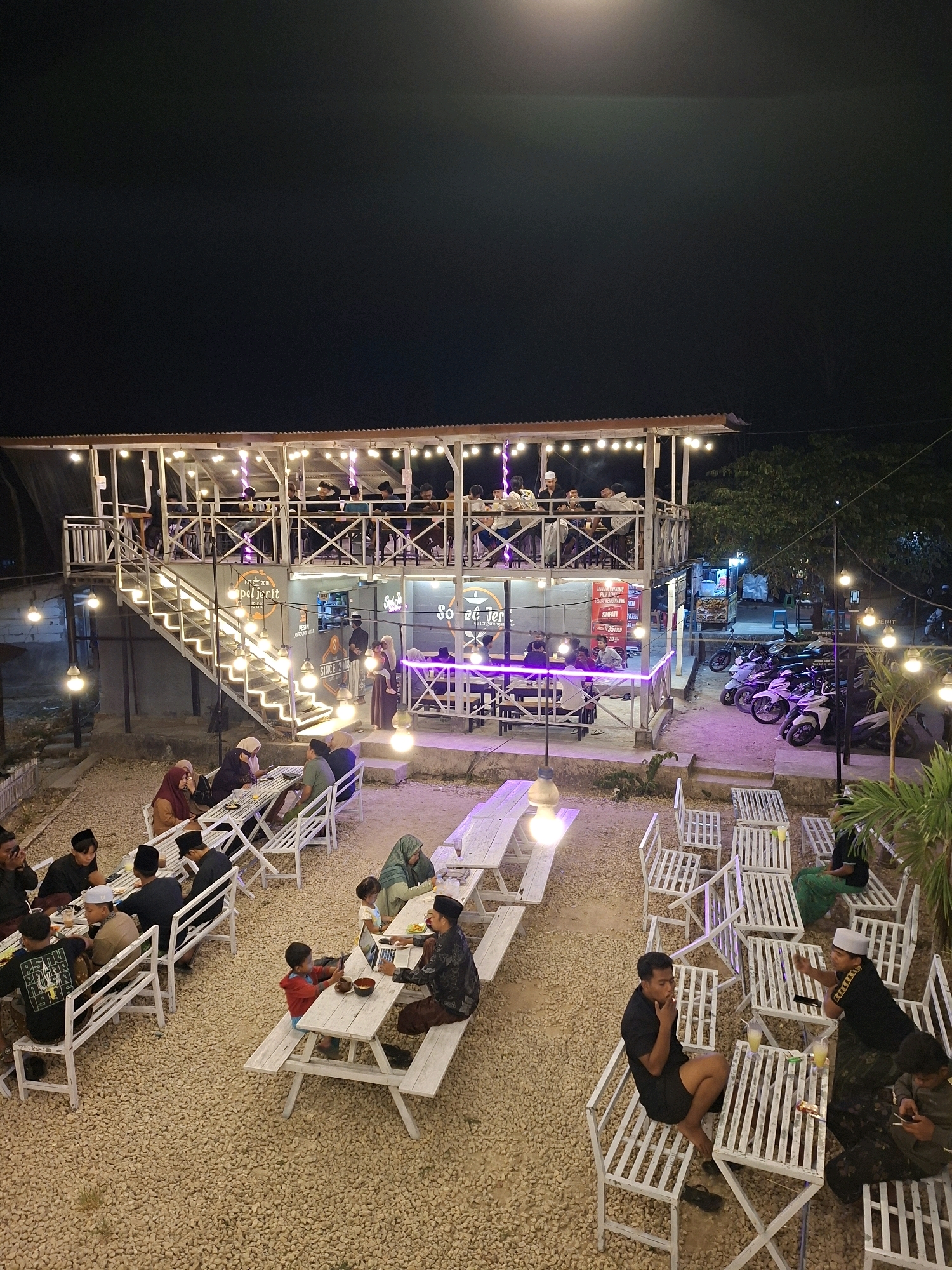 The image shows an outdoor dining area at night, likely a cafe or restaurant, located in Lantek Barat, Indonesia. The establishment has a two-story structure with an outdoor staircase leading to the upper level. The ground level features numerous white picnic tables and benches arranged on a gravel surface. String lights illuminate the area, creating a festive atmosphere.
Several groups of people are seated at the tables, engaged in dining and conversation. Some individuals are wearing traditional Indonesian attire, including peci hats and headscarves. The upper level also appears to be occupied, with people seated at tables and visible through the railing.
In the background, a row of motorcycles is parked, suggesting that customers arrive by vehicle. The overall scene depicts a lively social gathering and dining experience in a tropical, outdoor setting. The lighting and arrangement of tables suggest a place designed for communal outdoor dining.
