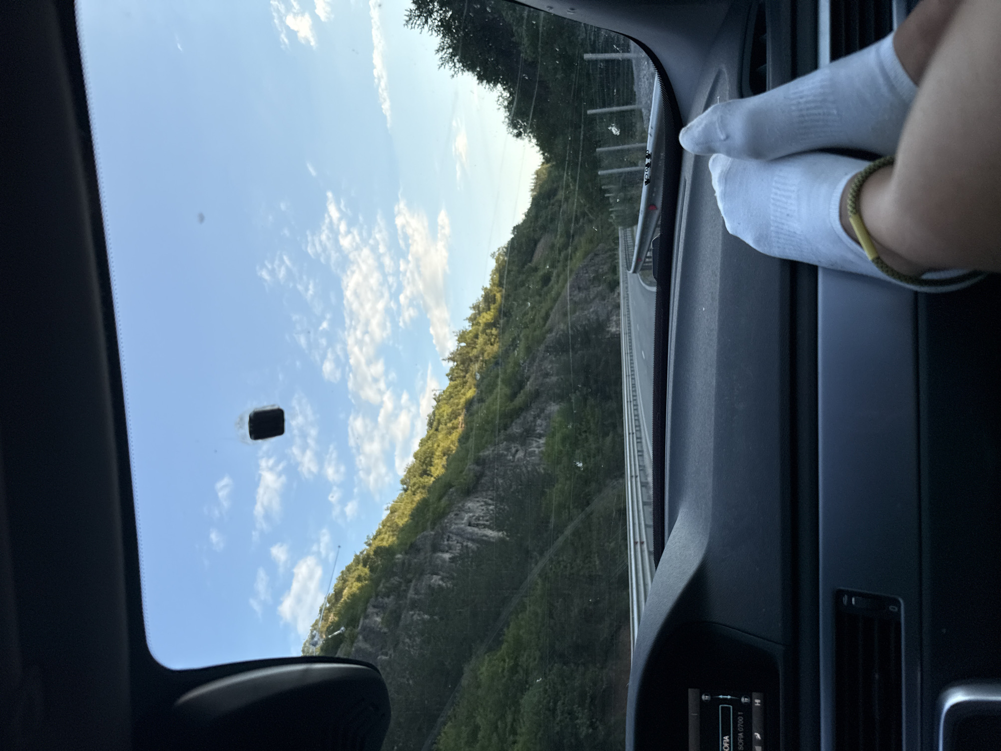 The image shows a view from inside a car, looking out through the windshield. The car's dashboard is visible in the foreground, with a small screen displaying "SOFIA" and "SOFIA 0700 1". Two pairs of feet, wearing white socks, are resting on the dashboard, one foot intertwined with the other, and a green bracelet is visible on one ankle. Outside the car, a road curves through a mountainous, forested landscape. The sun is shining, casting light on the green hills and creating some shadows. The sky is blue with scattered white clouds. The scene suggests a journey, possibly on a scenic route. The location is identified as Kalotina, Bulgaria, a border crossing town between Bulgaria and Serbia.