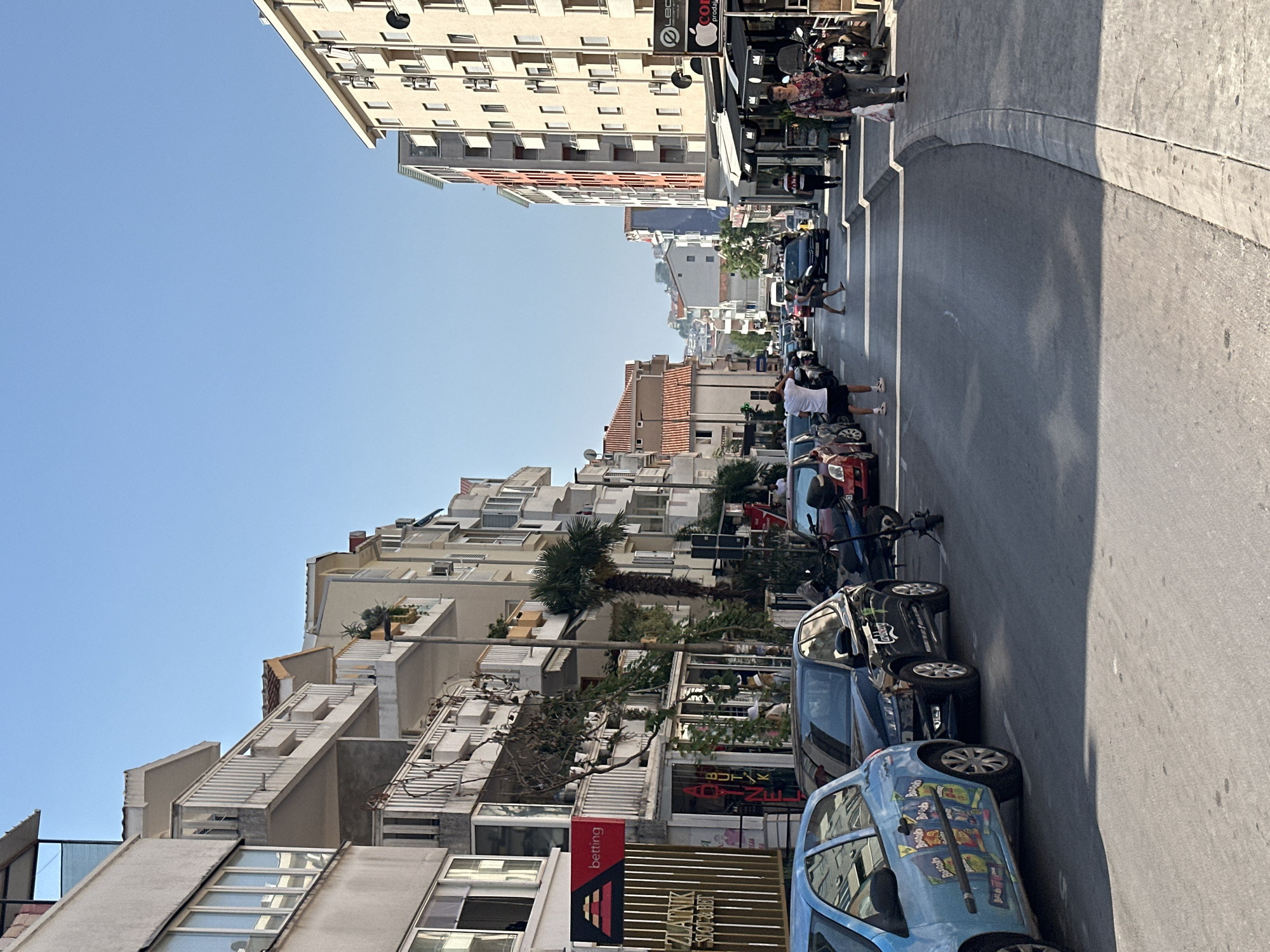 The image depicts a street scene in Budva, Montenegro, on a clear, sunny day. The street is lined with multi-story buildings featuring balconies and numerous windows. The architecture suggests a Mediterranean or Balkan influence. Several parked cars and a scooter are visible along the side of the road. Pedestrians are walking on the sidewalks and crossing the street. One man is standing in the road, and another is walking across a crosswalk. The buildings on the left have shops on the ground floor, with one sign reading "betting" and another "ZLATNIK SLOT & BET." On the right, there are awnings over storefronts. The sky is a vibrant blue, and the overall impression is of a busy, urban environment during daylight hours.