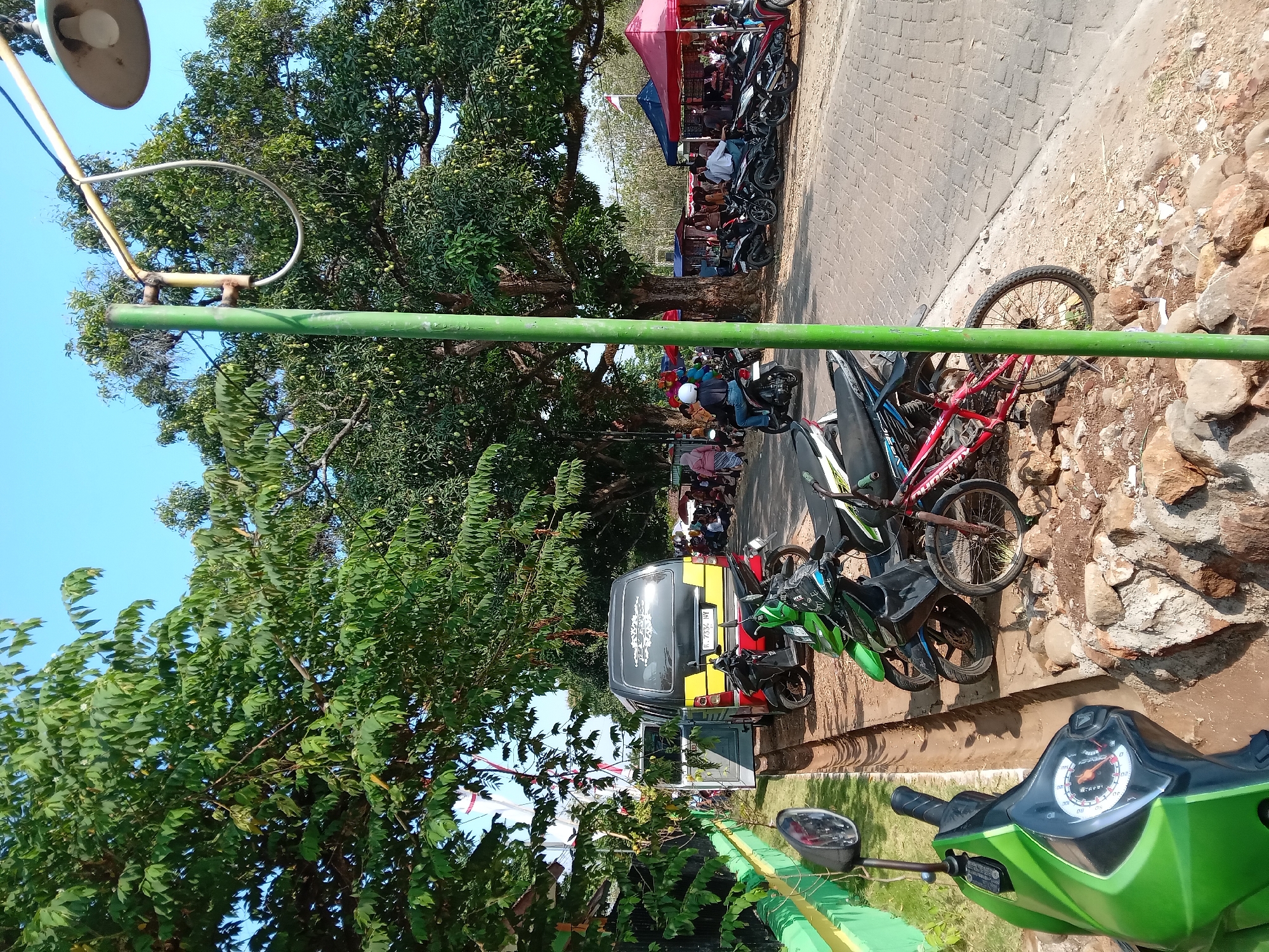 The image depicts an outdoor scene in Kedunggudel, Indonesia, characterized by a bright, sunny day and a clear blue sky. The foreground is dominated by the front of a green scooter, with its handlebars and instrument panel visible. To the right of the scooter, a narrow, dry ditch runs parallel to a paved pathway made of interlocking gray bricks.
Beyond the ditch and pathway, several motorcycles are parked. A black motorcycle and a red bicycle are situated closest to the foreground. Further back, more motorcycles are visible, interspersed with what appear to be market stalls or vendor tents. These tents are colorful, with a red and pink one prominently featured on the right side of the image.
A yellow and black van is parked on the pathway, its rear door open, revealing part of its interior. Behind the van and stalls, large, leafy trees with visible fruit, likely mangoes, create a shaded canopy. Several people are seen walking and standing around the market area, some on motorcycles and others browsing. A green lamppost stands near the center of the image, supporting a street lamp. The overall impression is of a bustling local gathering or market.