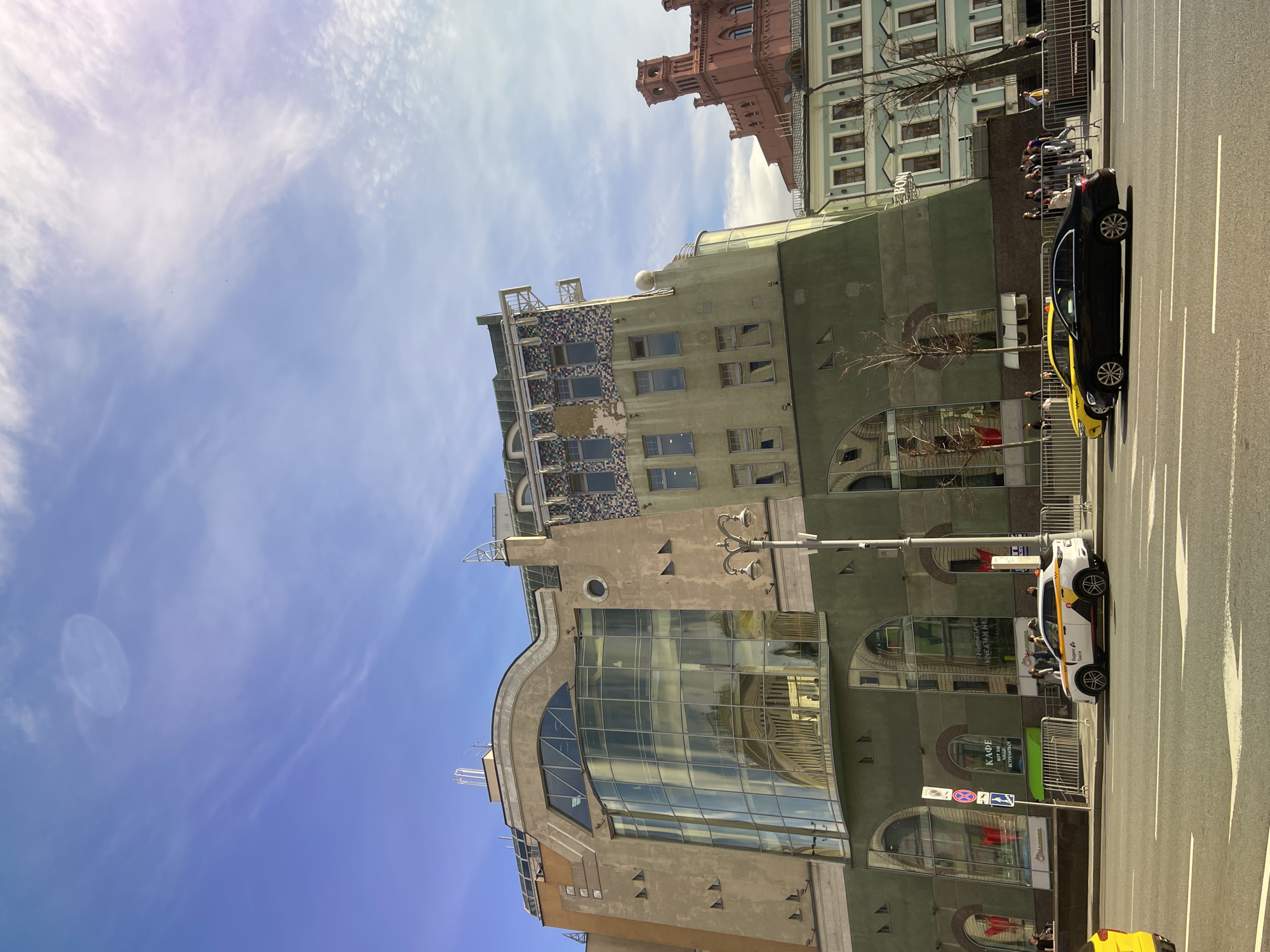 The image is a street scene in Moscow, Russia, showing a modern building with architectural elements, including a large glass facade and mosaic detailing on the upper floors. The building is light green and beige. Other buildings, including one in red brick, can be seen in the background. There are several people on the sidewalk behind metal barriers. Two cars, one black and yellow and one white with yellow stripes, are parked on the street next to the building. The sky is blue with scattered clouds.