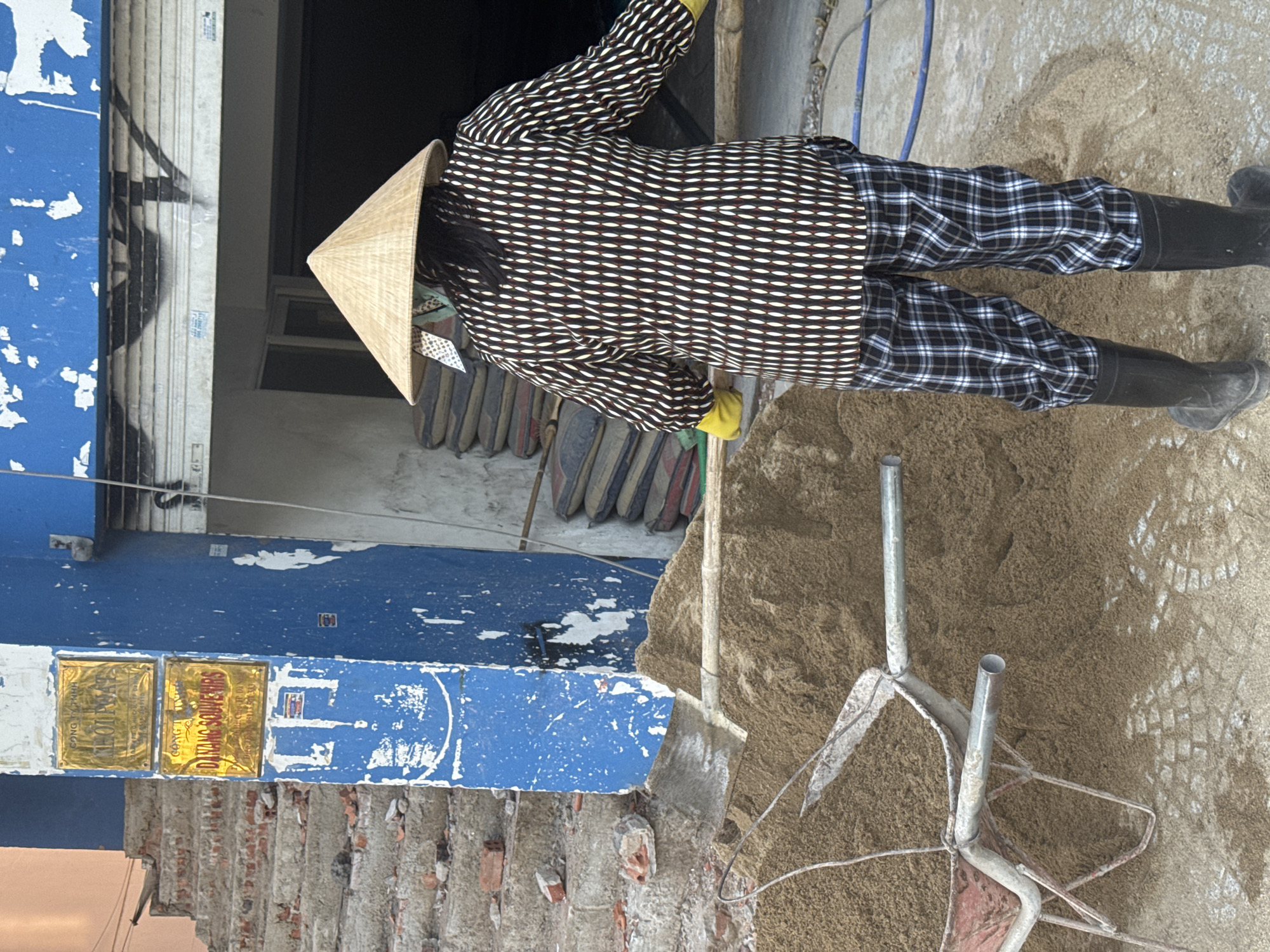 A person wearing a conical Asian hat, a patterned long-sleeved shirt, plaid pants, and black rubber boots shovels a pile of sand in front of a building in Đà Nẵng, Vietnam. The building's blue façade is peeling and features a sign that reads "DANANG SOUVENIRS." A damaged staircase is located nearby.