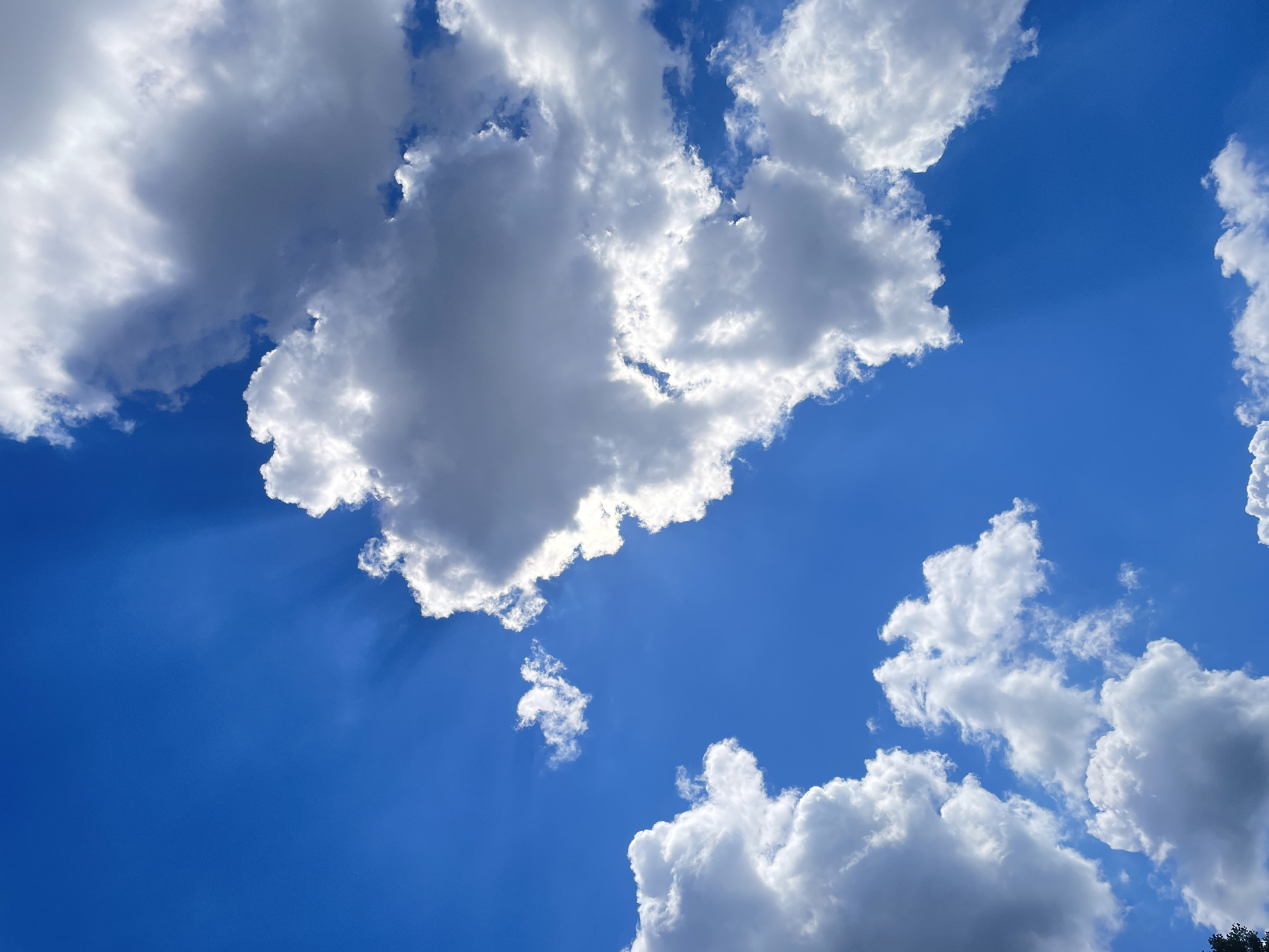 A bright blue sky is partially obscured by numerous cumulus clouds. Sunlight streams through gaps in the clouds, creating visible sunbeams. The clouds range in color from bright white to dark gray, indicating varying depths and densities. The image appears to be taken from a ground-level perspective looking upwards. No specific landmarks or indications of a city location are visible.