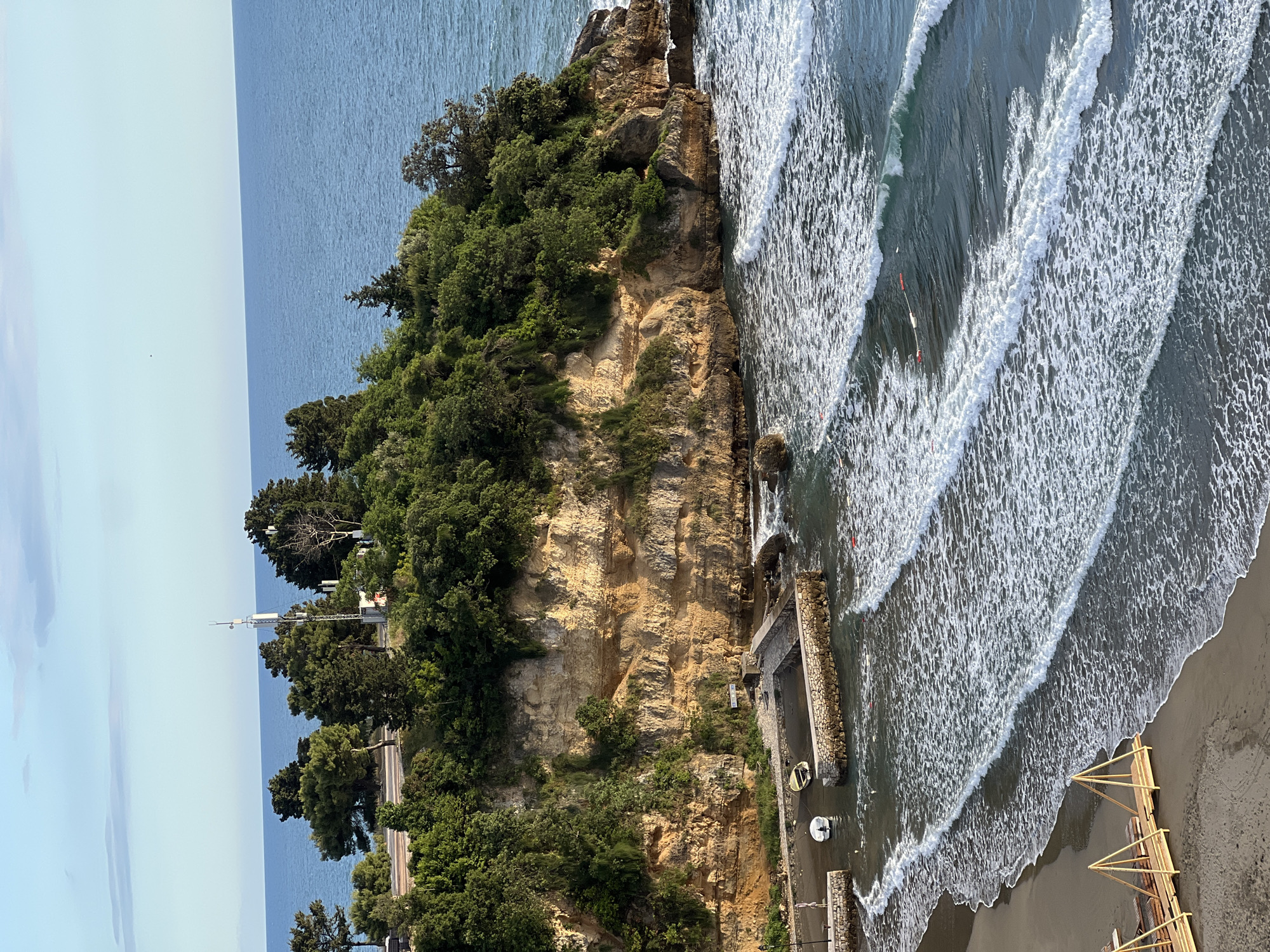 The image depicts a coastal scene in Ulcinj, Montenegro. A rocky cliff, heavily vegetated with trees, dominates the landscape. Below the cliff, a man-made stone pier extends into the sea, with small boats moored near it. Waves crash against the coastline. In the water, there are several individuals, likely swimmers or surfers. The beach is visible in the lower right of the frame, with wooden structures present. In the distance, a calm sea meets a light blue sky. Along the cliff, there is a road. Further in the distance, there appears to be a building with what appears to be antennas.