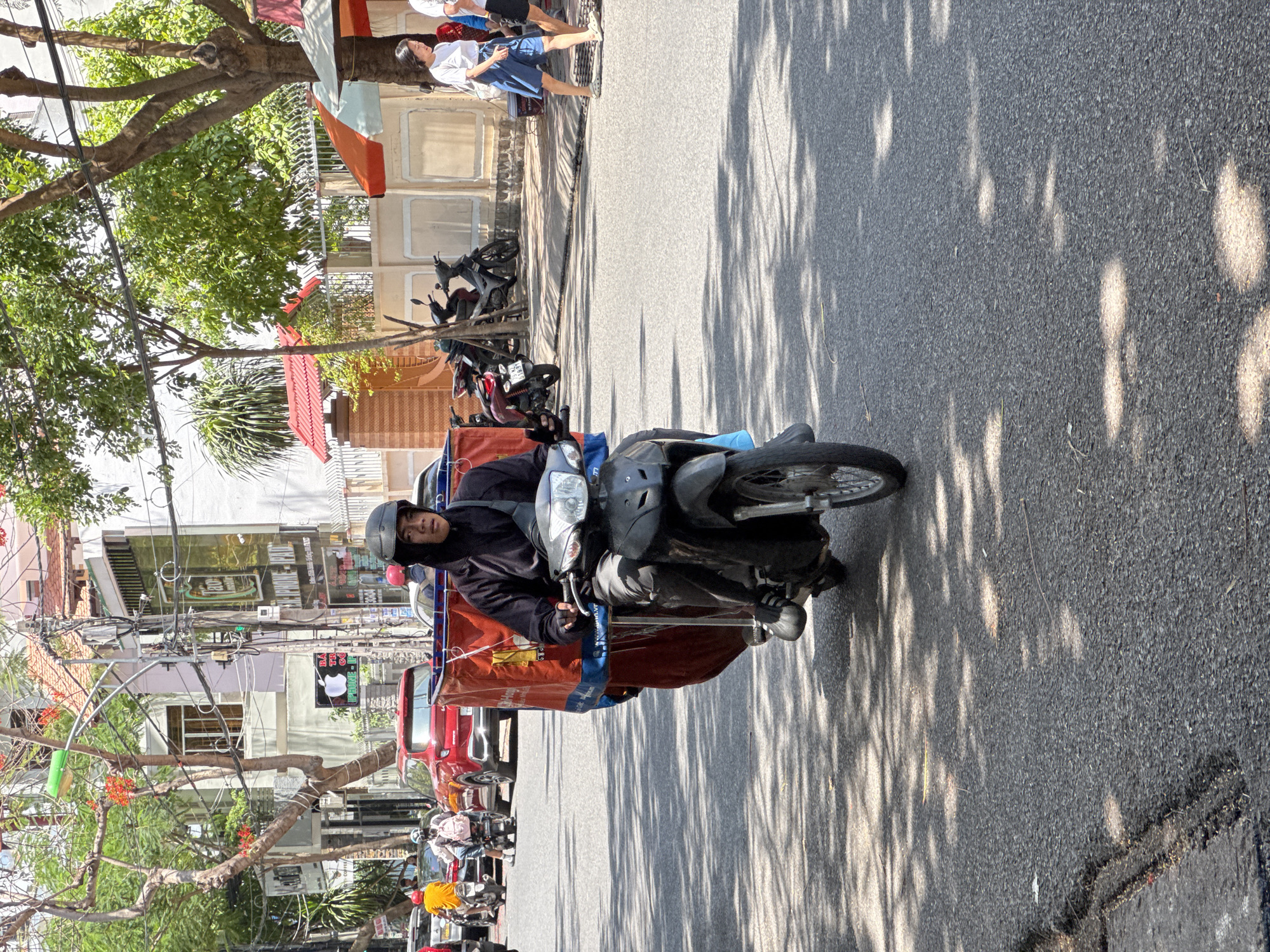 A male delivery driver wearing a black hooded jacket and a dark helmet rides a black motorcycle with a large, red insulated cargo box mounted on the rear. The motorcycle is in motion on a paved city street in Nha Trang, Vietnam, casting shadows from overhead trees onto the asphalt. In the background, other vehicles, including a red car, and pedestrians are visible on the sidewalk. Buildings with various signs, including one with an Apple logo, line the street. The scene captures a moment of daily life and commerce in a bustling urban environment.