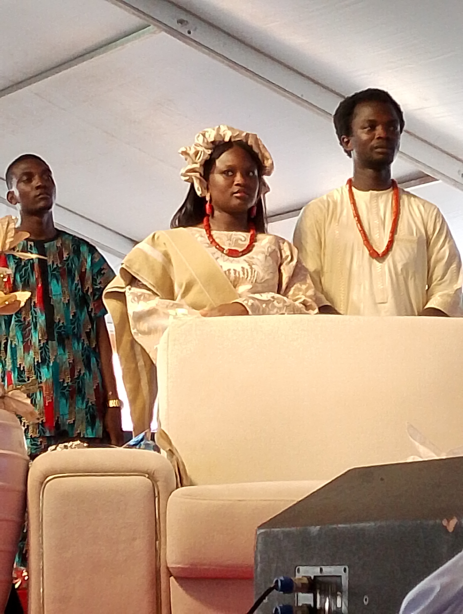 This full-length, slightly low-angle shot captures three individuals, two men and one woman, standing under what appears to be a large white event tent or temporary structure in Dushepe, Nigeria. The scene suggests a formal cultural event or ceremony.
The central figure is a woman, dressed in a light-colored, possibly cream or beige, patterned dress. She wears a matching decorative head wrap (gele) and striking red coral beads as a necklace and earrings, indicating cultural significance. Her expression is calm and composed, looking forward.
To her right stands a man, also looking forward with a neutral expression. He is attired in a light-colored, possibly white or cream, traditional long-sleeved tunic (agbada or similar style) and a prominent red coral bead necklace, mirroring the woman's accessory.
On the woman's left, the third individual, a man, wears a vibrant, intricately patterned long-sleeved shirt in shades of teal, red, and black, possibly a dashiki. He has a gold-colored bracelet on his right wrist and looks slightly upward with a neutral gaze.
In the foreground, part of a light-colored, plush upholstered sofa or armchair is visible, with its armrest and back supporting the woman's arm. To the far left, a tall, light-colored decorative vase or urn is partially seen. On the lower right, a dark grey speaker or piece of sound equipment with visible cables is present. The ceiling structure of the tent, with its metal supports, is clearly visible above the subjects, illuminated by bright, diffused light, suggesting daytime or strong artificial lighting. The traditional attire and coral beads strongly suggest a Nigerian cultural celebration, possibly a wedding or engagement ceremony.