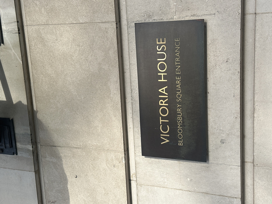 A dark, rectangular, metallic sign with gold lettering is affixed to a light-colored, textured stone or concrete building facade. The sign reads "VICTORIA HOUSE" in prominent uppercase letters, with "BLOOMSBURY SQUARE ENTRANCE" in smaller uppercase letters below. The facade exhibits horizontal mortar lines and vertical joints between panels. Shadows from an unseen overhead structure are cast onto the upper wall section and to the left of the sign. Faint discolored streaks are visible on the facade above and around the sign. The scene depicts an entrance sign for Victoria House, specifically the Bloomsbury Square entrance, located in London, United Kingdom.