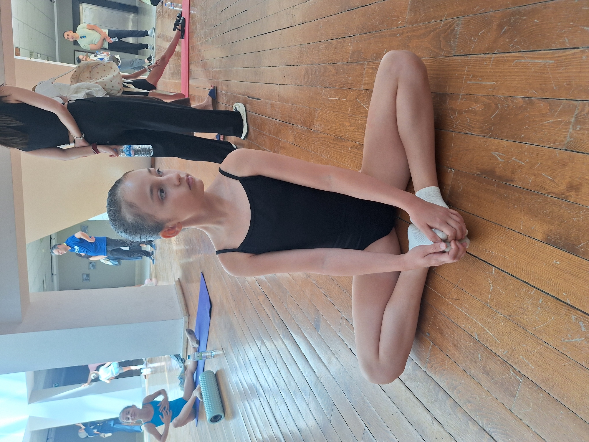 A young girl in a black leotard and white socks is shown from the chest down, sitting on a wooden floor in a dance studio. She is holding her feet, with her knees bent and legs in a butterfly stretch pose. Her gaze is directed upwards and to the left. In the background, other people, including a woman in black pants and a white top, and a man in a light green t-shirt and dark pants, are visible, as well as a pink yoga mat and a water bottle. Further in the background, a mirrored wall reflects more of the studio and several other individuals, some of whom are also engaged in what appears to be a dance or gymnastics class. The lighting suggests it is daytime, and the overall atmosphere is one of activity and instruction. The location is Çankaya, Turkey.