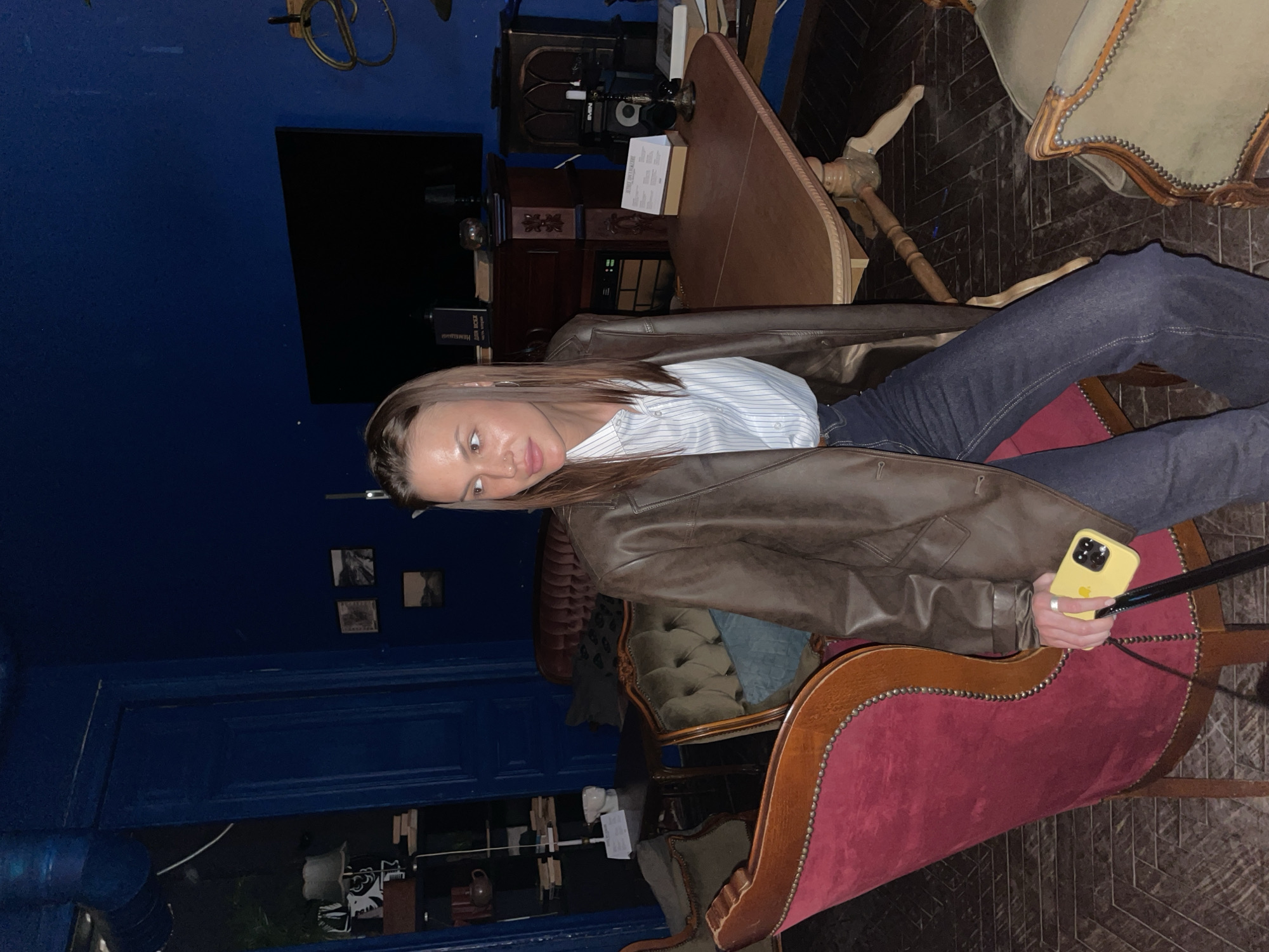 A female is seated on a red velvet chair, holding a yellow iPhone. She wears a brown leather jacket over a blue and white striped shirt and dark jeans. The location appears to be indoors, with a blue wall and antique furniture. A dark wooden table is visible in the background. The city and region are unknown.