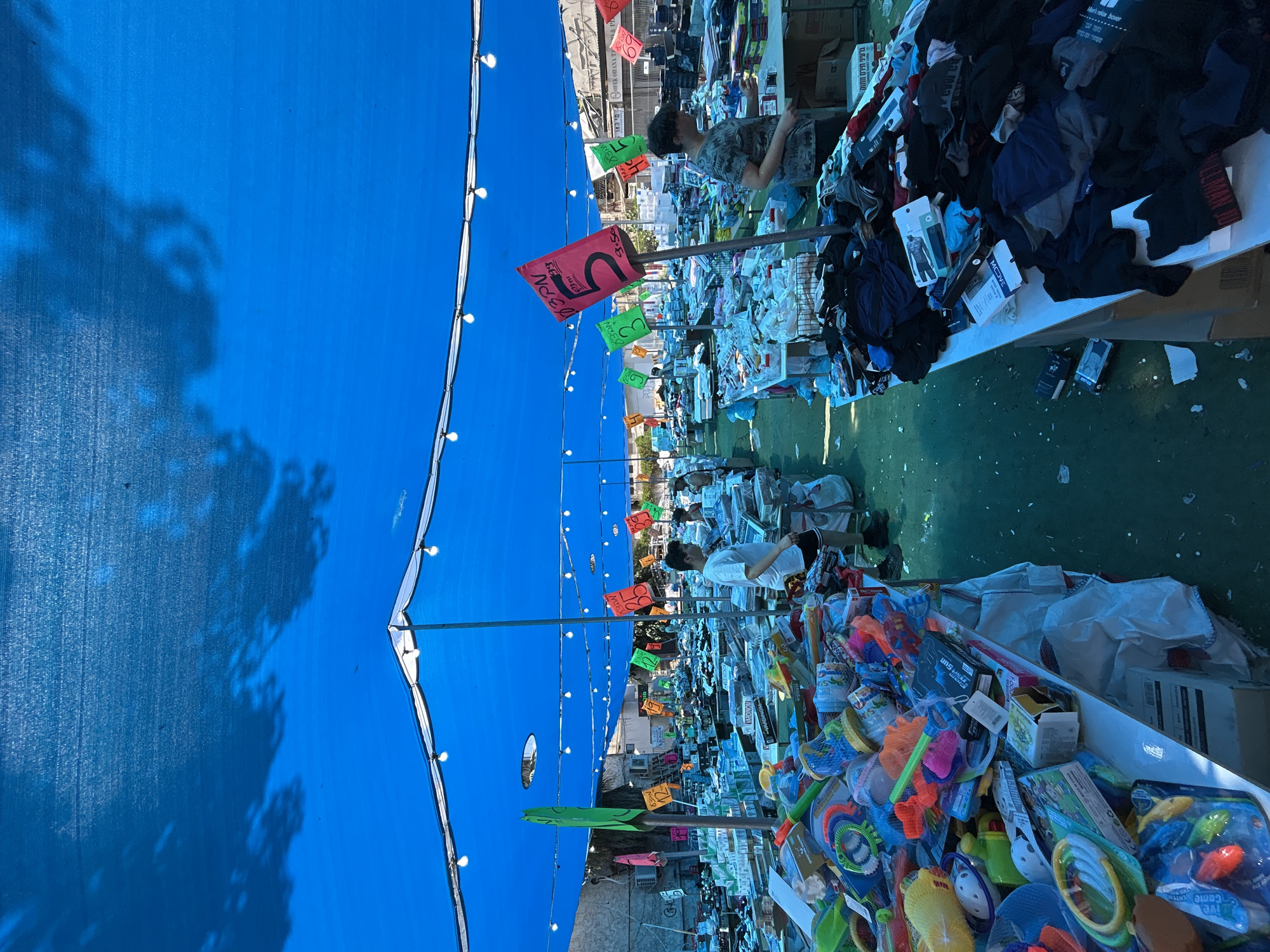 The image depicts an outdoor market under a large blue tarp, likely in Rishon LeZion, Israel, due to the Hebrew signage. The market is set up with rows of tables displaying merchandise, primarily clothing and toys. In the foreground, to the left, a large pile of colorful toys is visible, including buckets, sand molds, and water guns. To the right, tables are covered with folded clothing, mostly dark-colored items like t-shirts and socks, with some items laid out in stacks. Several people are present in the market. In the middle ground, a young man in a white t-shirt and black shorts is browsing, while further back, another person in a camouflage t-shirt is looking at the clothing. Other individuals can be seen in the background, also appearing to shop. Colorful signs with prices in Hebrew are hung on poles throughout the market, indicating prices such as "5", "3", and "12". The ground is covered in a green matting, and there is some litter scattered around. The overall scene is one of a busy, casual retail environment.