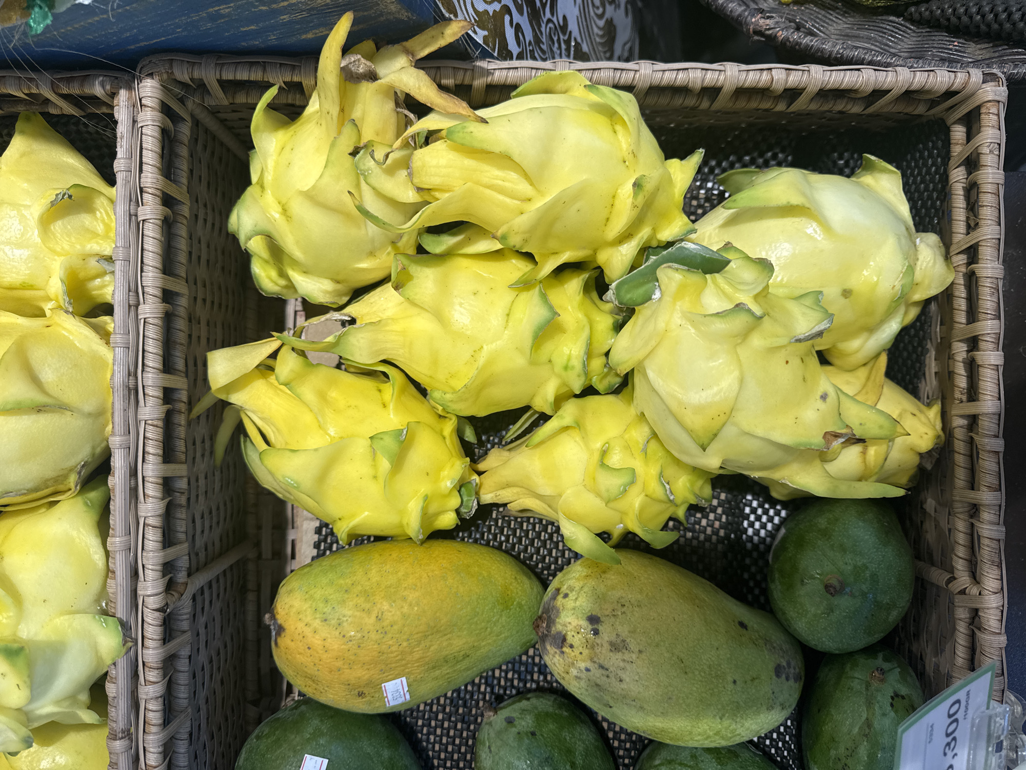 A close-up shot shows two woven baskets containing fruit, likely taken in Kuta Selatan, Indonesia, judging by the currency on a price tag. The baskets contain yellow dragon fruit and green mangoes. The fruits are displayed in a retail setting, likely a market or grocery store.