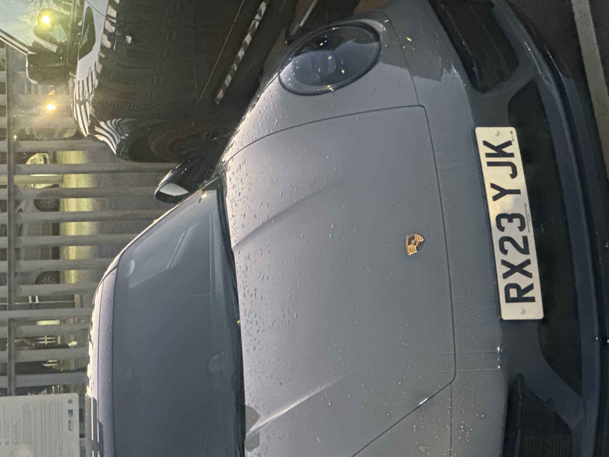 The image displays the front-left section of a grey Porsche 911, likely a 992 generation, parked in an outdoor or semi-enclosed commercial area. The vehicle's hood, windshield, and roof are covered in numerous small water droplets, indicating recent precipitation. Faint vertical streaks are discernible on the hood, originating above the license plate. The front United Kingdom license plate is clearly visible, reading "RX23 YJK," with "Porsche Centre Tonbridge" inscribed in smaller text below the main characters. To the right of the Porsche, a dark-colored vehicle, consistent with a modern Land Rover Defender, is partially visible, its front-right headlight reflecting brightly. In the background, a light-colored metal fence or barrier runs horizontally, separating the foreground from an indistinct area containing blurred forms and artificial light sources. No individuals are visible, and the scene is static.
Regarding the location in the city of Sevenoaks, United Kingdom: The license plate's inscription "Porsche Centre Tonbridge" identifies a car dealership located approximately 5-6 miles southeast of Sevenoaks. While the precise location within Sevenoaks cannot be definitively ascertained from the visual evidence, the scene's environment is consistent with a vehicle dealership or commercial parking facility in the broader Kent area.