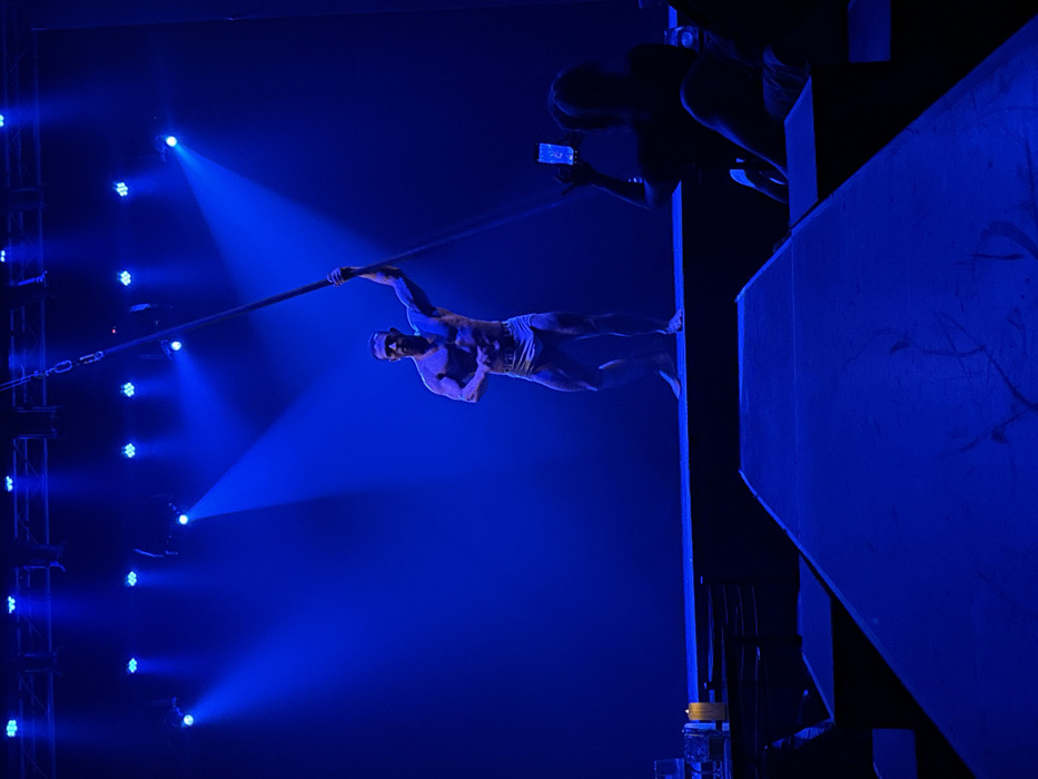 A blue-lit circus performance features a muscular man balancing horizontally on a tightrope, holding a pole for stability. A second person leans from above a platform, taking a photo with a phone. The lighting rig with spotlights is visible on the left.