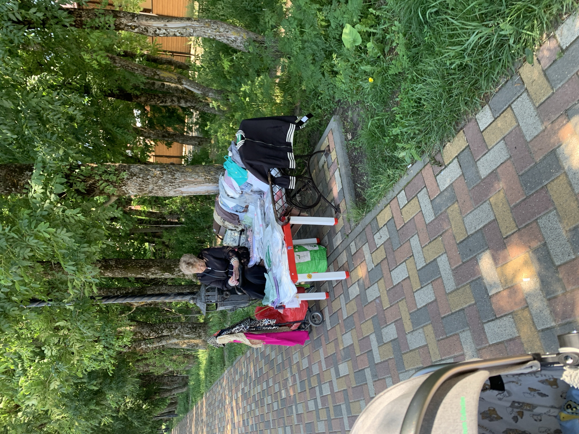An elderly woman sits at a table laden with folded clothes on a sidewalk lined with trees and a grassy verge. She is looking at her phone. A small trolley with more clothes and bags is next to the table. In the foreground, the edge of a stroller is visible. The pavement is made of multi-colored rectangular bricks. The environment suggests a park or public walkway in the city of Stavropol, Russia.