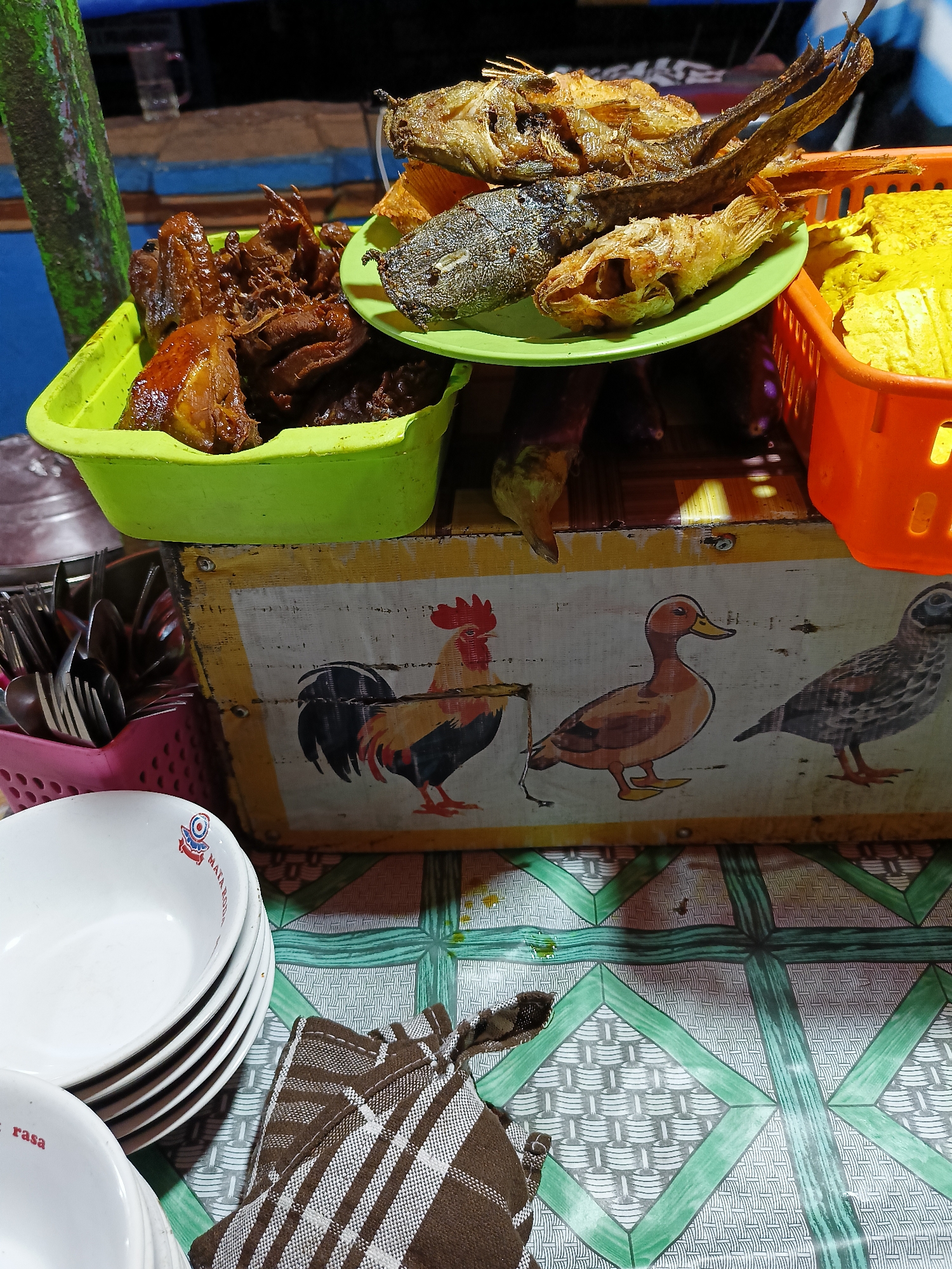 An outdoor food stall displays various fried dishes and utensils. In the foreground, a stack of white bowls with red and blue logos rests on a patterned tablecloth. Beside the bowls, a folded brown and white checkered cloth lies on the tablecloth. In the mid-ground, a wooden chest is decorated with painted images of farm animals: a rooster on the left, a duck in the center, and a bird on the right. To the left of the chest, a pink basket holds an assortment of silverware. Above the chest, a light green plastic container holds pieces of dark, saucy fried food, possibly chicken. A green plate, stacked on top of the container, holds a variety of fried fish, including what appears to be catfish and smaller whole fish. To the right of the chest, an orange plastic basket contains bright yellow fried items, likely tempeh or tofu. The background is blurred, showing dark, possibly wooden structures and a glimpse of a glass. The overall scene depicts a food vendor's setup, likely in a public area of Kebondalem Kidul, Indonesia, at night, indicated by the artificial lighting.