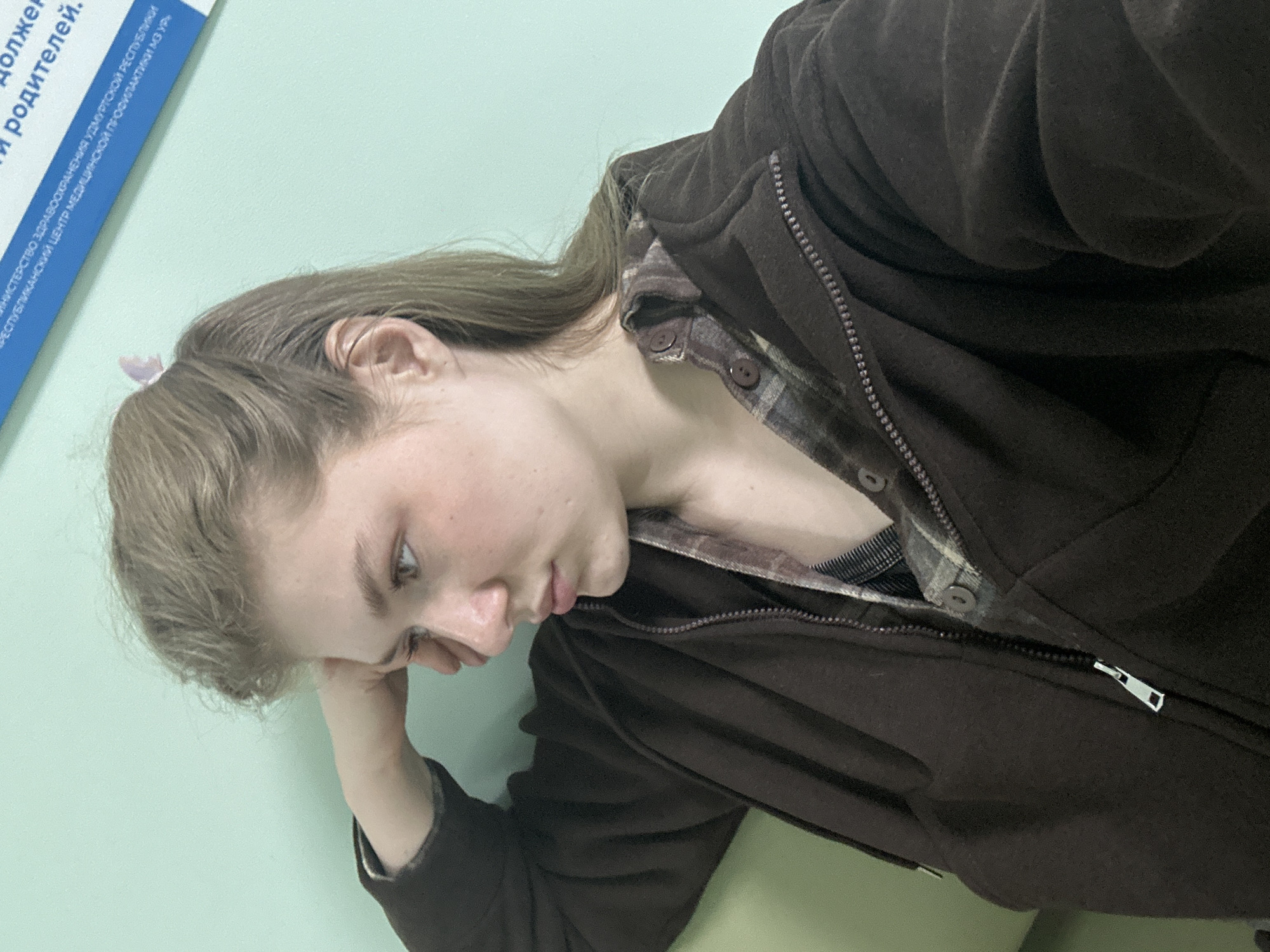 The image displays a young female, fair-skinned with light brown hair, seated or leaning against a plain light teal-colored wall. She is depicted in profile, facing left, with a pensive or passive expression. Her right hand is raised, supporting her head with her palm against her temple. A small, light-colored butterfly clip is visible in her hair, which is partially pulled back. She is wearing a dark brown, zippered hoodie over an unbuttoned plaid shirt featuring brown, grey, and white checks, with a dark undergarment visible. A silver-colored zipper pull is visible on the hoodie. In the upper right background, a blue rectangular sign is partially visible, bearing white Cyrillic text that identifies the location as a "Ministry of Health of the Udmurt Republic Republican Center for Medical Prevention." The scene is located within a medical or public health facility in Мунайка, Udmurt Republic, Russia.