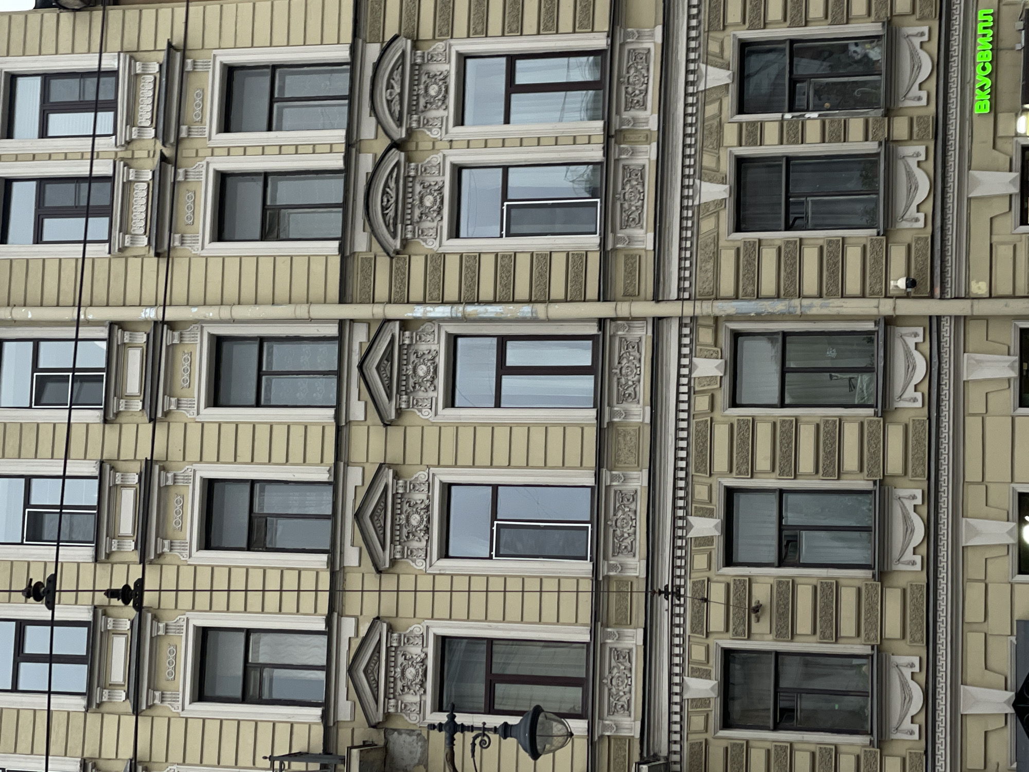The photograph depicts the facade of a building in Saint Petersburg, Russia. The architectural style features ornate detailing, with rows of windows set within elaborately decorated frames. The building's exterior is predominantly beige or light yellow. There are visible utility lines and a streetlamp in the foreground. The composition is vertically oriented, focusing on the building's facade. A sign in Russian is partially visible.