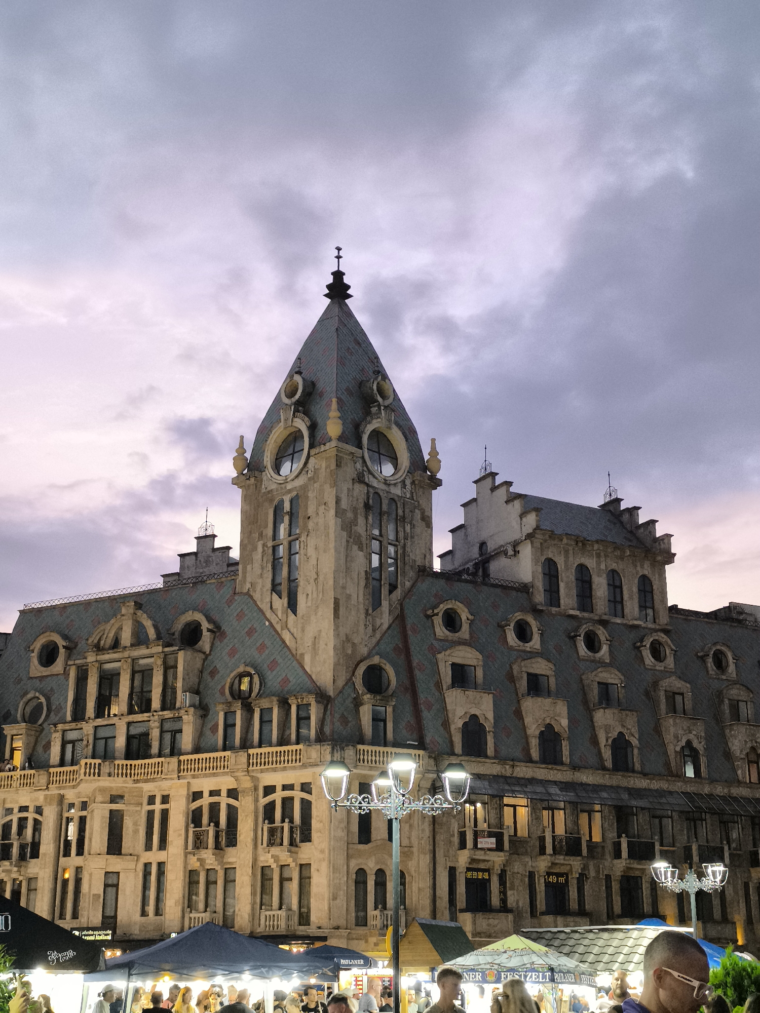 The image shows the Batumi Astronomical Clock, located in Batumi, Georgia. The clock tower, part of a larger historic building, is visible against a twilight sky with scattered clouds. The building features ornate architecture with a steeply pitched, multi-colored roof and numerous windows. In the foreground, a street-level market or fair is set up with tents and lights, and several people are visible browsing or standing around. A street lamp with multiple lights stands in the middle ground, illuminating the scene.