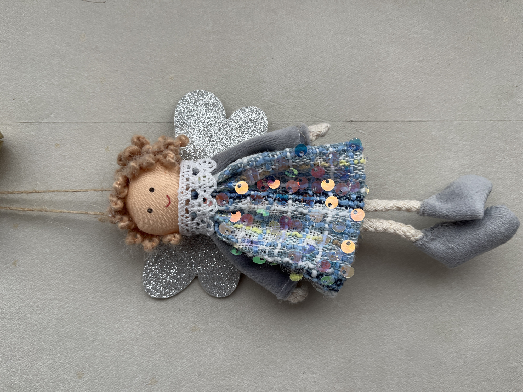 This is a close-up, eye-level shot of a handmade fabric angel ornament lying on a textured, light gray surface. The angel is positioned horizontally from left to right in the frame.
The angel's head is a round, peachy-colored fabric with two black dots for eyes and a small red curve for a mouth. It has light brown, curly yarn hair. Silver glittery heart-shaped wings are attached to its back. The angel wears a blue, white, and black textured fabric dress adorned with scattered colorful sequins, primarily in pink, blue, and gold tones. A white lace trim encircles the neck of the dress. The angel has gray fabric arms, one of which is visible and appears to be tucked into the dress. Its legs are made of braided twine, and it has two gray, rounded, sock-like feet. A thin piece of twine is visible extending from the left side of the ornament, suggesting it is part of a garland or string.
The background is a plain, light gray material with a subtle textured surface, possibly paper or fabric. There are a few faint, darker specks on the background surface. The lighting is even, and there are no strong shadows, indicating diffused light. The overall mood is whimsical and decorative. There is no visible text in the image.