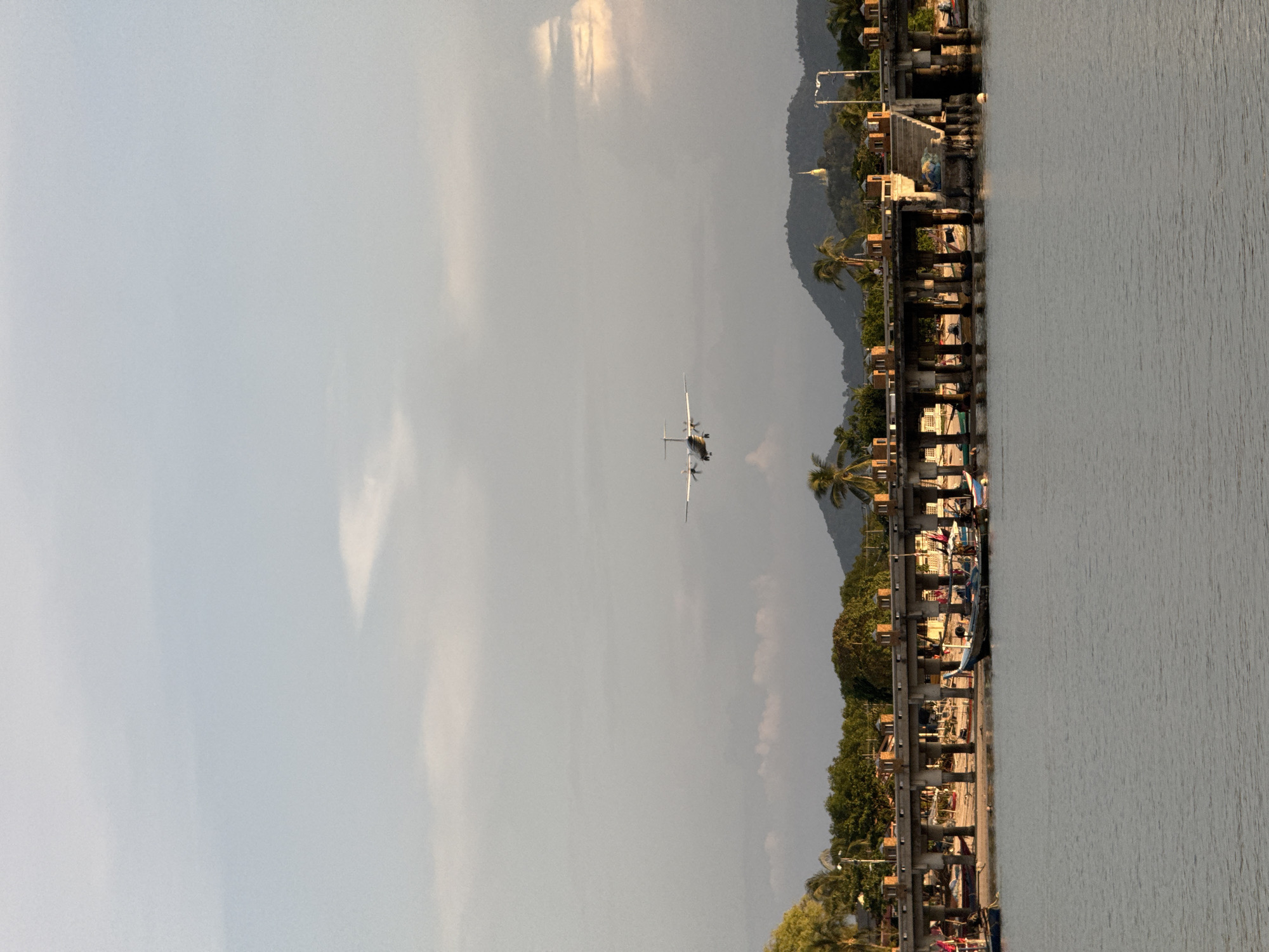 An airplane is flying in the sky over a coastal scene in the city of Ko Samui District, Thailand. The airplane, a twin-engine turboprop, is flying from left to right. Below the airplane is a long, elevated concrete pier with pillars. Several small fishing boats are docked or beached along the shore beneath the pier. The shore is lined with trees, including palm trees, and buildings are visible in the background. In the distance, hills rise, topped with a white stupa or temple. The sky is partly cloudy with soft sunlight. The water in the foreground is calm.