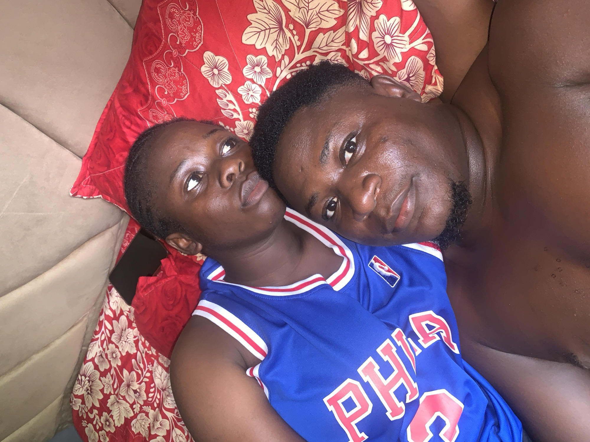The image shows a close-up, overhead view of two individuals, a woman and a man, lying down. The woman is on the left, her head resting on a red pillow with a floral pattern. She is looking upwards with her eyes open. She is wearing a blue basketball jersey with red and white trim. The man is on the right, his head resting next to the woman's. He is looking towards the camera with his eyes open and a slight smile. He appears to be shirtless. The setting appears to be a bed or sofa, with a beige fabric surface visible on the left. The lighting suggests it is indoors, possibly during the day or evening. The overall mood seems intimate and relaxed. The visible text on the basketball jersey includes "PHILA" in large red letters and an NBA logo. The provided location context is Akwanga, Nigeria.