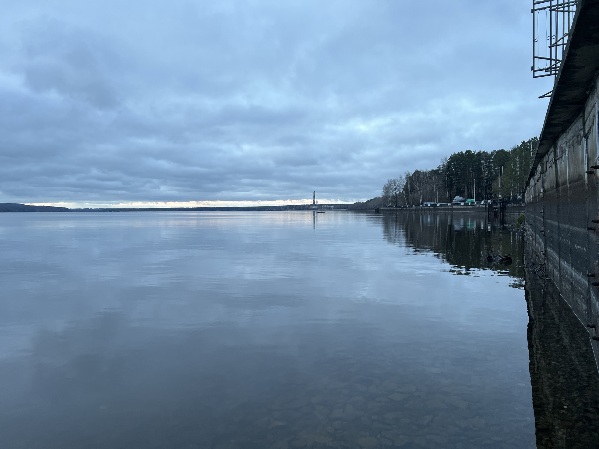 The image depicts a body of water, likely a reservoir or lake, near the city of Chaikovsky, Russia. The sky is overcast with a layer of gray clouds, reflecting on the water's calm surface. The horizon line reveals a distant treeline with a tall structure, potentially a factory chimney or tower, slightly off-center. In the foreground, on the right, there's a concrete retaining wall with metal structures attached, partially extending into the water. The water near the wall is shallow and clear enough to see the rocks at the bottom. A forested area stretches along the right edge of the image, adjacent to the wall. There are hints of green and white structures or vehicles alongside the treeline. The overall impression is one of a calm, industrial waterfront landscape under a somber sky.