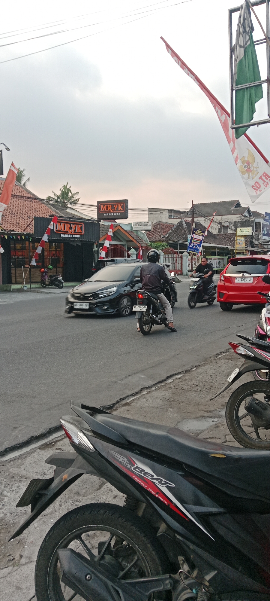 The image depicts a street scene in Maguwoharjo, Indonesia, under an overcast sky. In the foreground, the rear section of a black and red scooter is visible, with its tire and part of the body. The middle ground shows a traffic intersection. Several vehicles are present: a black car is turning left, a motorcyclist in a black jacket and helmet is riding a motorcycle in the same direction as the car, and a red car is stopped in the right lane. Behind the red car, another motorcycle is partially visible.
On the left side of the street, a barbershop named "MR.YK BARBERSHOP" is identifiable by its signage. The shop has a modern facade with black siding and orange and white accents, decorated with Indonesian flags. Further down the street, other buildings and structures are visible, including houses with tiled roofs and some advertisements.
The environment appears to be a typical urban or suburban street with paved roads and sidewalks. Power lines are visible crisscrossing the sky. The overall impression is one of everyday activity in a bustling area of Maguwoharjo.