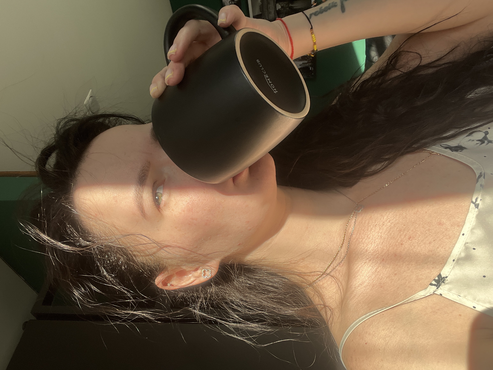 A woman is holding a black mug labeled "HOMECLUB" up to her face. She has long dark hair, light skin, and is wearing a silver necklace and a white top with a pattern. A red string bracelet and a bracelet with yellow beads are visible on her wrist. The background appears to be a dimly lit room with a window casting sunlight. There is no information to confirm this photo was taken in Kazan, Russia.