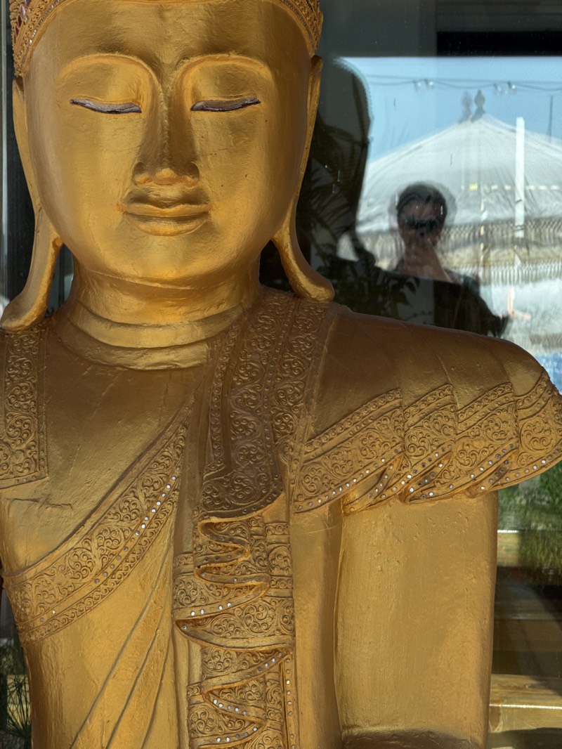 A close-up, eye-level shot captures a golden statue, likely a Buddha or Bodhisattva, dominating the left and center of the frame. The statue displays a serene and meditative expression with closed eyes and a subtle smile. Its head is adorned with an ornate, multi-layered crown, and its robes are intricately patterned with raised golden designs and small, reflective silver accents or jewels along the edges and folds. The surface of the statue has a warm, metallic sheen.
The statue is situated indoors, placed in front of a large glass window or partition. This glass reflects an outdoor scene and a person. A distinct reflection of a person, presumably the photographer, is visible in the upper right quadrant of the glass, wearing sunglasses and a dark top. Behind and around this reflection, a large white, possibly tent-like or domed structure is clearly seen against a light blue sky, along with hints of green foliage.
The bright, natural illumination indicates that the photograph was taken during daytime, likely under sunny weather. The overall scene suggests a blend of cultural art within an indoor setting that connects to an outdoor environment. The location context for this image is Sant Antoni de Portmany, Spain. There is no visible text in the media file.