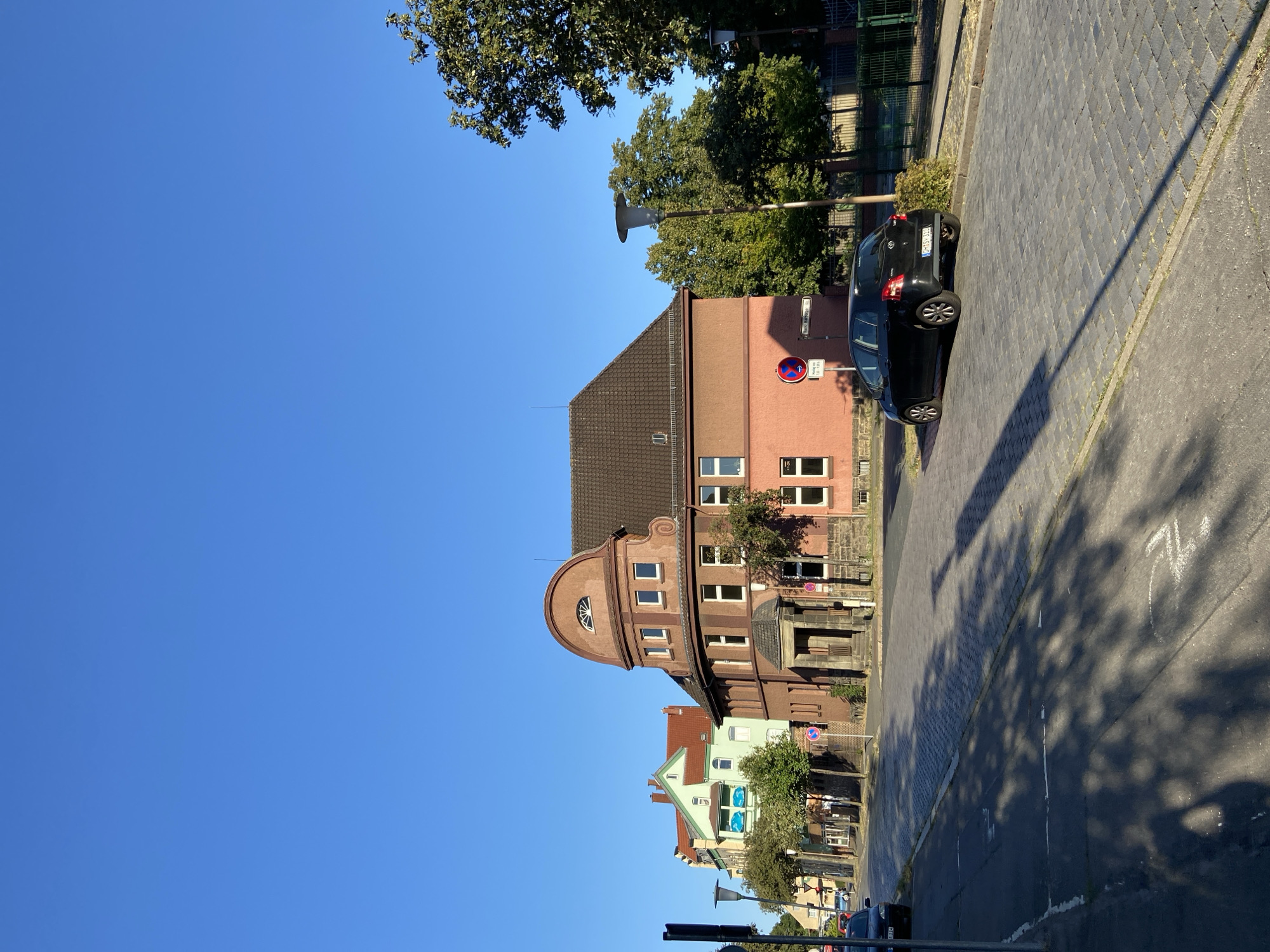 The image shows a street scene in front of several buildings. The sky is clear and blue. In the foreground, a paved road with cobblestone sections is visible, with shadows of trees cast across it. A black car is parked on the right side of the road. To the left, a sidewalk with a lamppost and a signpost is present.
Several buildings line the street. The building closest to the foreground is a multi-story structure with a red-orange facade. It features multiple windows, a prominent arched section at the top with a small window, and a covered entrance with stone steps. To the left of this building is another building with a light green facade, a red tiled roof, and a balcony with blue railings. Further down the street, more buildings with red roofs are visible.
Trees with green foliage are scattered throughout the scene, some with shadows falling on the road. There are various street signs and traffic signs visible, including a red and white circular sign with an arrow and a horizontal bar, and a rectangular sign with text. The environment appears to be a residential or mixed-use area within a city.