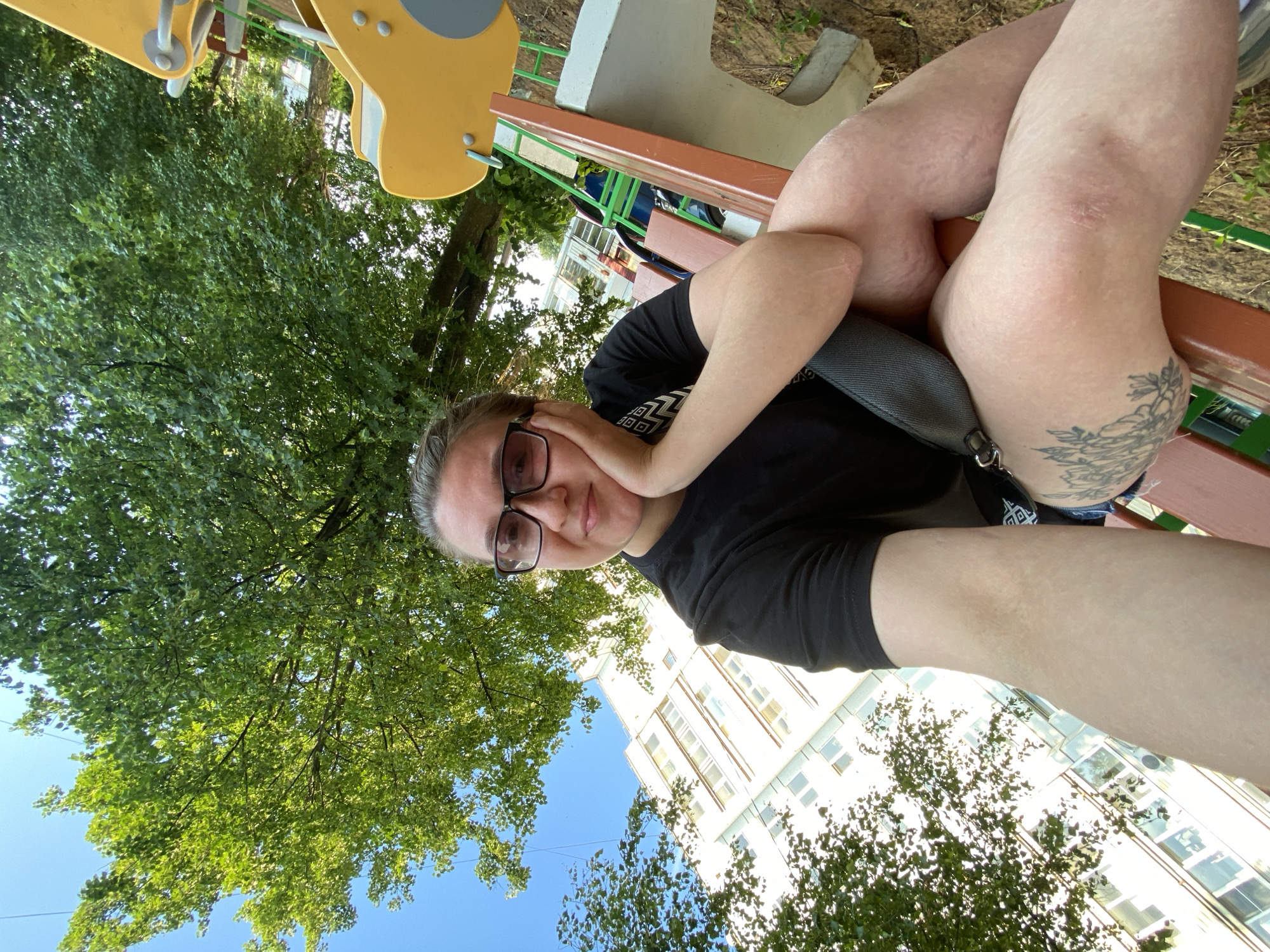 A woman with glasses, wearing a black t-shirt and denim shorts, is sitting on a park bench outdoors. She has a tattoo on her left thigh. The bench has a brown wooden top and a white concrete base. The background shows a residential building, a large green tree, and a bright blue sky. Parts of a yellow playground structure are visible in the foreground. The scene appears to be in Chaikovsky, Russia, likely a residential area with a playground.