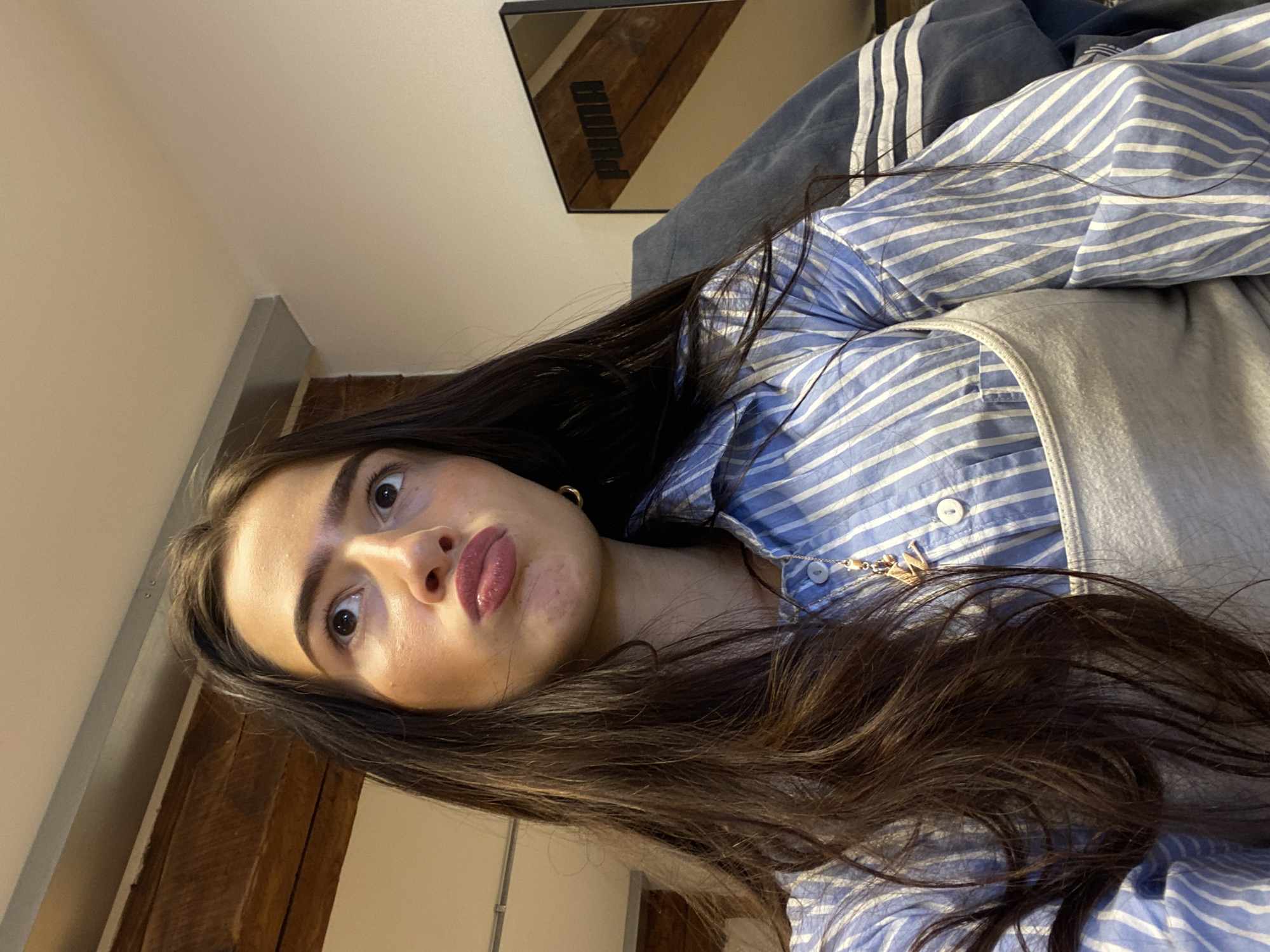 A woman with long, dark brown hair is pictured from a slightly low angle. She has fair skin and is wearing a blue and white striped shirt over a grey tank top. A gold necklace is visible around her neck. The background includes a wall, what appears to be wooden paneling, and a mirror with the word "PUMA" reflected in it. Other articles of clothing can be seen as well. The location is not identifiable as London.
