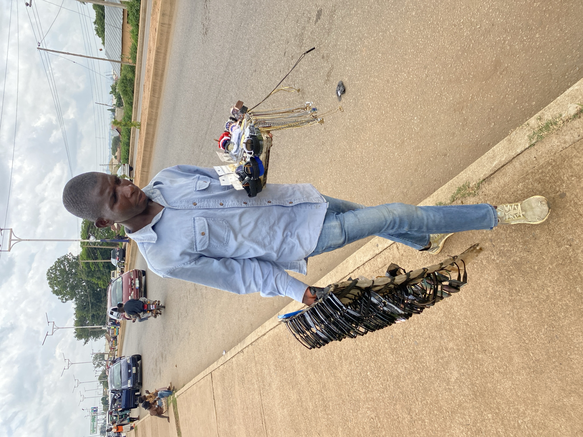 A young man, appearing to be in his late teens or early twenties, walks along a paved sidewalk beside a busy road in Bukuru, Nigeria. He is wearing a light blue long-sleeved collared shirt and faded blue jeans, with grey athletic shoes. He is carrying a tray in his right hand filled with an assortment of items, including several wristwatches of various designs and colors, as well as numerous necklaces and other jewelry. In his left hand, he holds a large bundle of sunglasses, stacked vertically. The road behind him is filled with traffic, including cars, motorcycles carrying passengers, and a bus. Several people are visible on the sidewalk and road in the background, some sitting and others walking. The environment appears to be a commercial or semi-urban area, with buildings and vegetation visible further in the distance under a cloudy sky.