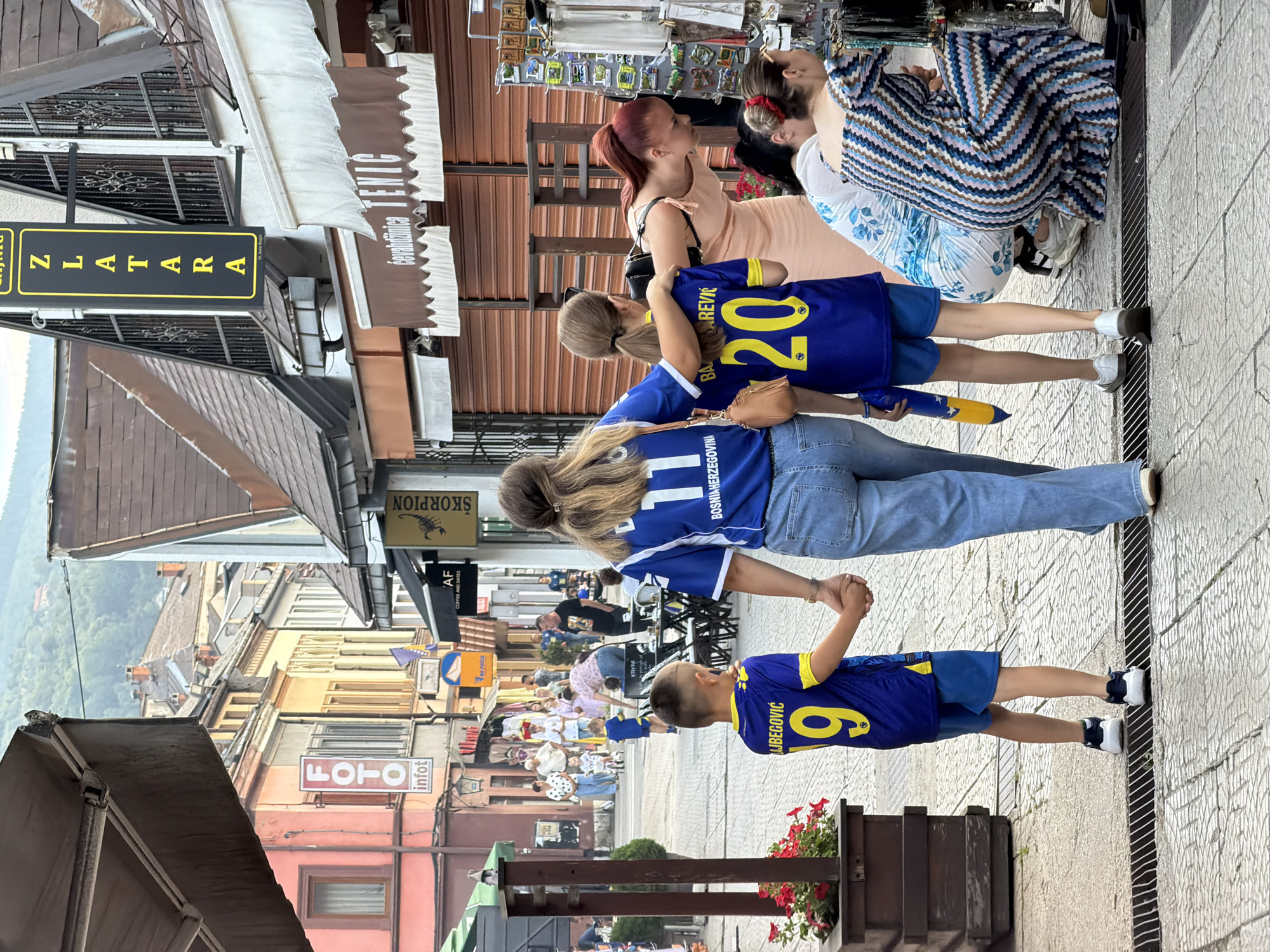 This is a photograph taken on a cobblestone street in Travnik, Bosnia and Herzegovina, likely in the old town area. The scene depicts a bustling pedestrian thoroughfare lined with buildings featuring traditional architecture. On the left, a building with a pink facade has signage for "FOTO" and "infot." Further down the street, other buildings display various shop signs including "SKORPION" and "CEVABDZINICA TENIC." The overall atmosphere suggests a commercial and tourist-friendly environment.
In the foreground, a family is walking away from the camera. A woman wearing a blue jersey with "11 BOSNIA AND HERZEGOVINA" on the back and blue jeans walks hand-in-hand with a young boy also in a blue jersey with the number "9" and the name "LAJBEGOVIC" on the back. Beside them walks a girl, also in a blue jersey with the number "20" and the name "BAJPAREVIC" on the back, with her arm around the girl in front. The girl is holding a yellow and blue inflatable object.
To the right, two women are browsing a stall displaying various items, possibly souvenirs like magnets. One woman, with red hair tied in a ponytail, wears a light peach-colored dress. The other woman is wearing a patterned blue and white dress.
In the background, more people are visible, some walking, some seated at outdoor tables of a cafe. The street is paved with cobblestones, and a drain grate is present in the foreground. The buildings in the background suggest a hilly landscape, typical of the region. The lighting indicates it is daytime, with overcast or diffused sunlight.
