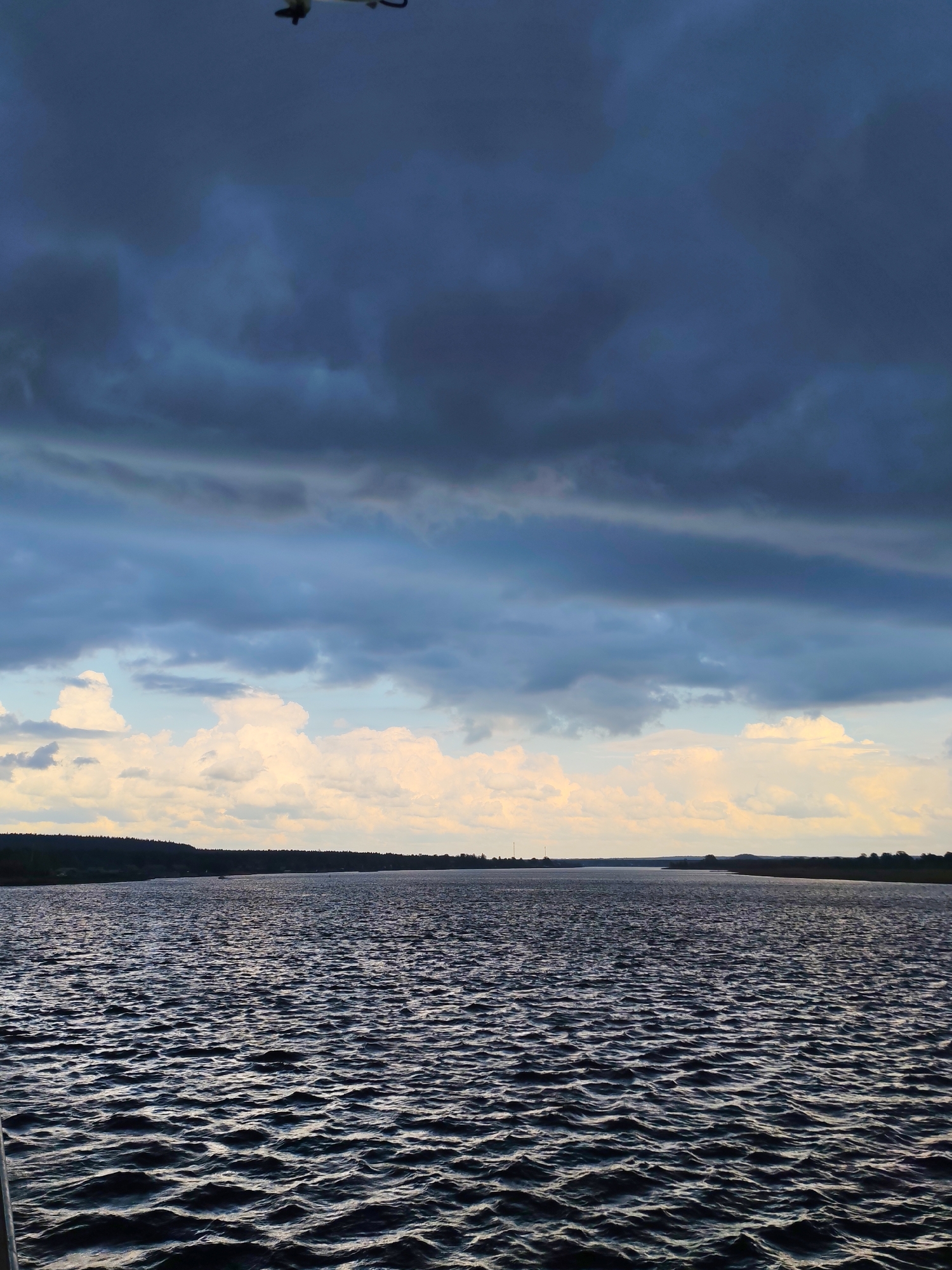 The image depicts a large body of water, possibly a river or lake, with moderate ripples on the surface. The foreground shows the water up close, with darker tones reflecting the overcast sky.
In the background, a thin strip of land is visible, likely the opposite shore, covered with trees. The sky is dominated by heavy, dark storm clouds, except for a strip of lighter, partially clear sky with white and yellow clouds just above the land.
There is a plane in the top center of the image.
The image is taken during daylight, but the lighting is subdued due to the heavy cloud cover. The geographical location is unspecified.