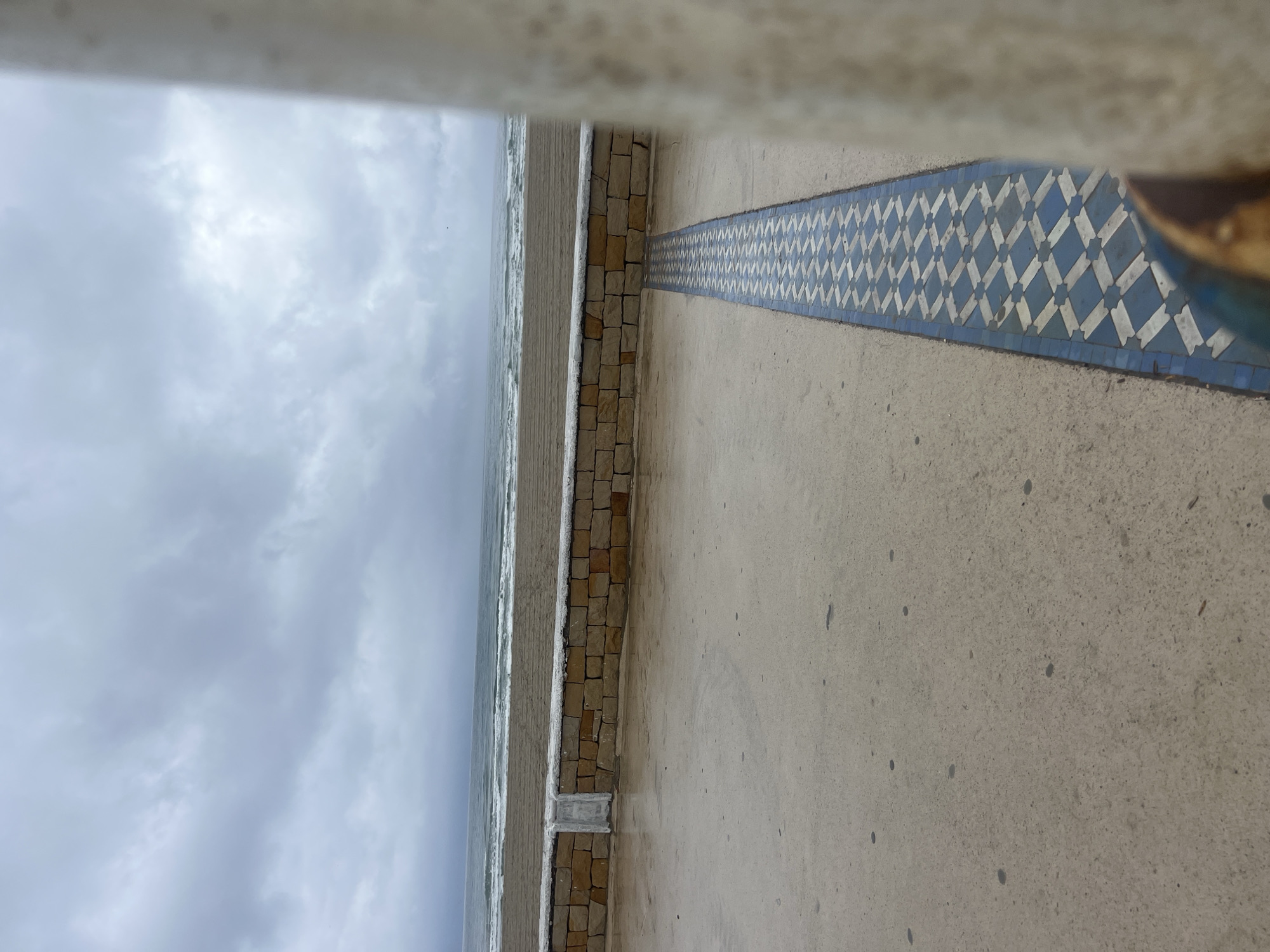 This landscape image captures a view from a seaside promenade in Martil, Morocco, looking towards the ocean on an overcast day. The upper portion of the frame is dominated by a cloudy, light grey sky, suggesting diffused light and possibly a cool temperature. Below the horizon, the greyish-blue expanse of the ocean features visible white-capped waves breaking near the shore.
In the foreground, a concrete promenade stretches across the width of the image. A low wall, constructed from small, rectangular blocks of light brown and tan stone, separates the promenade from the beach and ocean. Running parallel to this stone wall on the concrete surface is a distinctive decorative strip of blue and white mosaic tiles. The tiles are arranged in a repeating geometric pattern of interlocking diamonds or rhombuses. The concrete surface of the promenade exhibits small dark speckles.
A blurred, light-colored architectural element, possibly a railing or an overhang, is visible across the very top of the frame, suggesting the vantage point is partially obscured. Another blurred, brownish object appears at the far right edge, potentially another part of the surrounding structure. There are no people visible in the scene, and no specific activity is taking place. The overall impression is one of a calm, perhaps somber, day by the sea. No text is visible in the image.