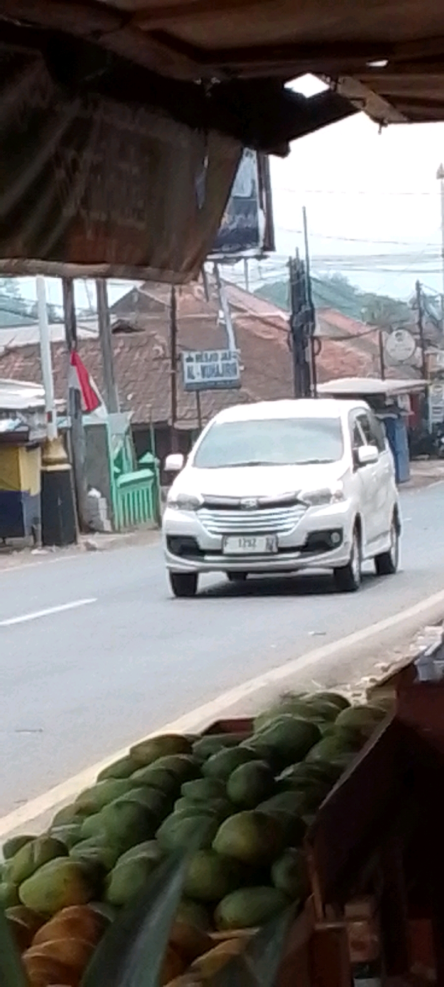 In the city of Cilaku, Indonesia, a white minivan with license plate F 1752 is traveling on a paved road. In the background, a sign reads "MASJID JAMI AL-YUHAIRIN," indicating a mosque. To the left of the mosque, a small green fence and a pole with a red and white flag are visible. In the foreground, a display of green fruits, possibly mangoes, is arranged on a stand. The scene appears to be a roadside market, with a stall of produce in the foreground and a mosque in the background. The sky is overcast, suggesting a cloudy day.