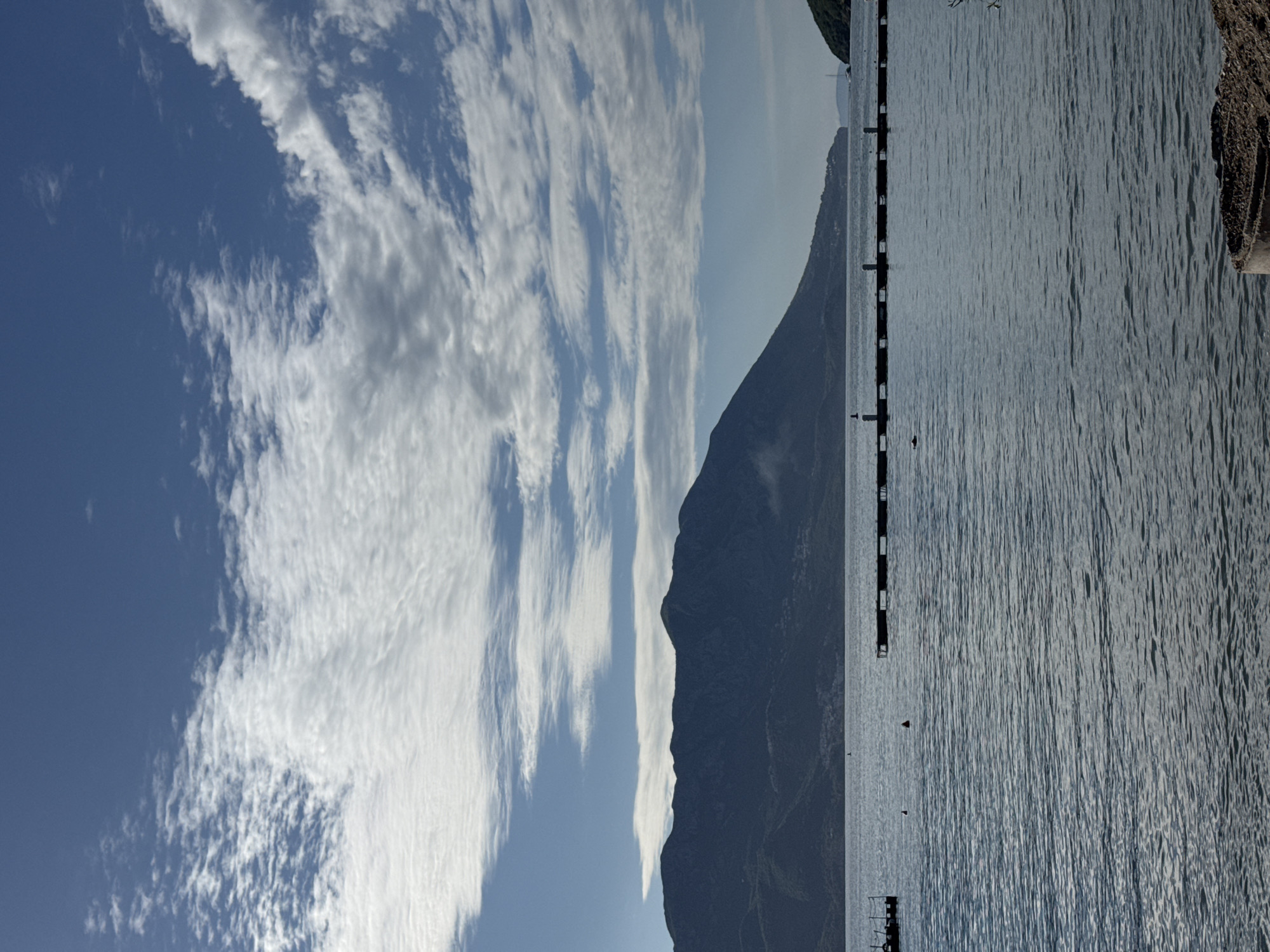 The scene depicts a landscape in Budva, Montenegro. A large, dark mountain rises in the center, separating the sky from the water. The sky is blue with streaks of white clouds. The water is calm, with horizontal lines suggesting gentle waves. A pier extends into the water, marked by regular posts with small platforms. Vegetation appears on a raised bank at the edge of the water.