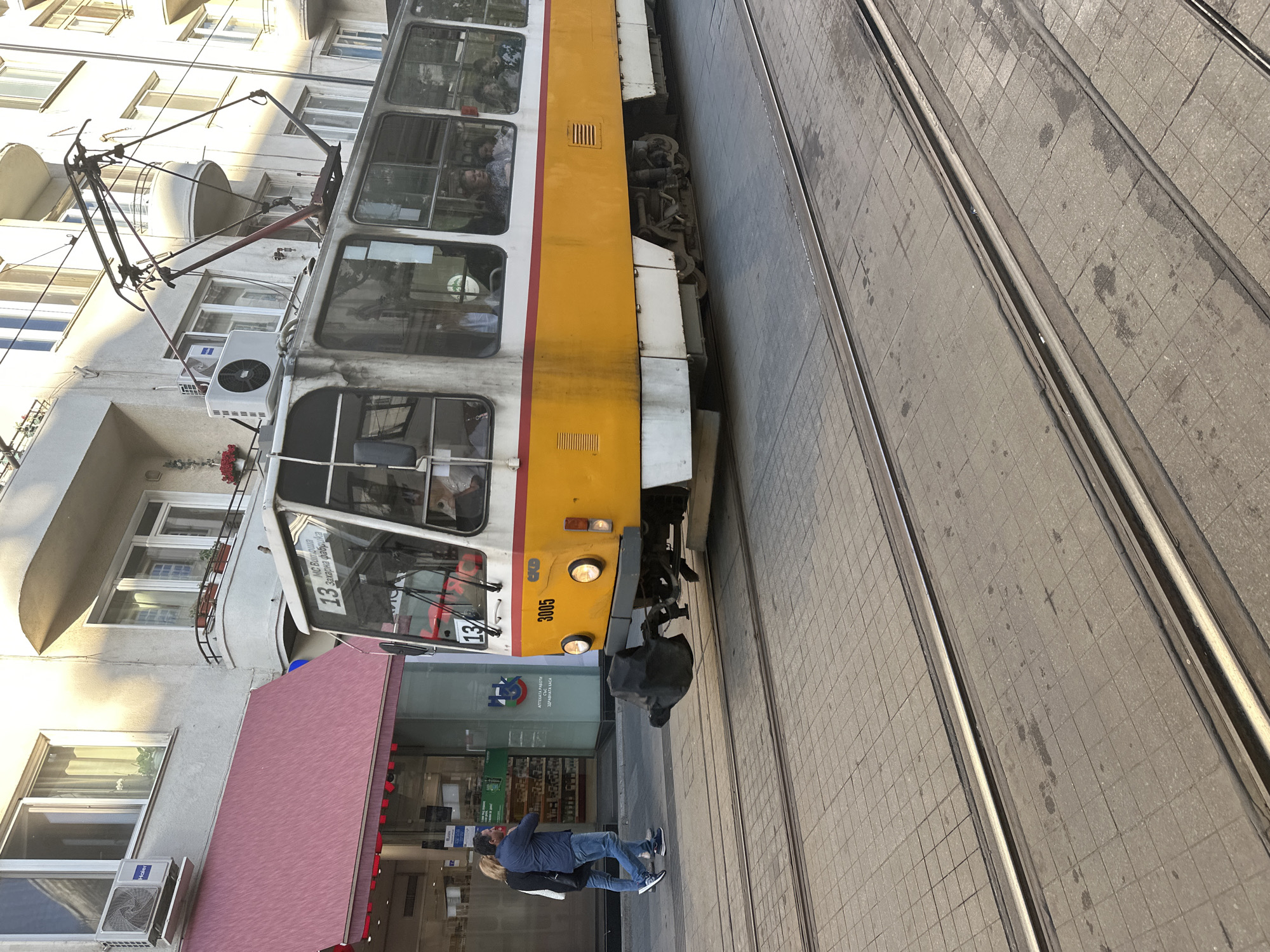 The image depicts a yellow and white tram, number 13 and marked "3005," traveling on rails in Sofia, Bulgaria. The tram is next to a building with a red awning, and a man and a woman are standing on the sidewalk next to the building. The tram's pantograph is visible on top, and the tracks run parallel to the building. The road is paved with cobblestones.